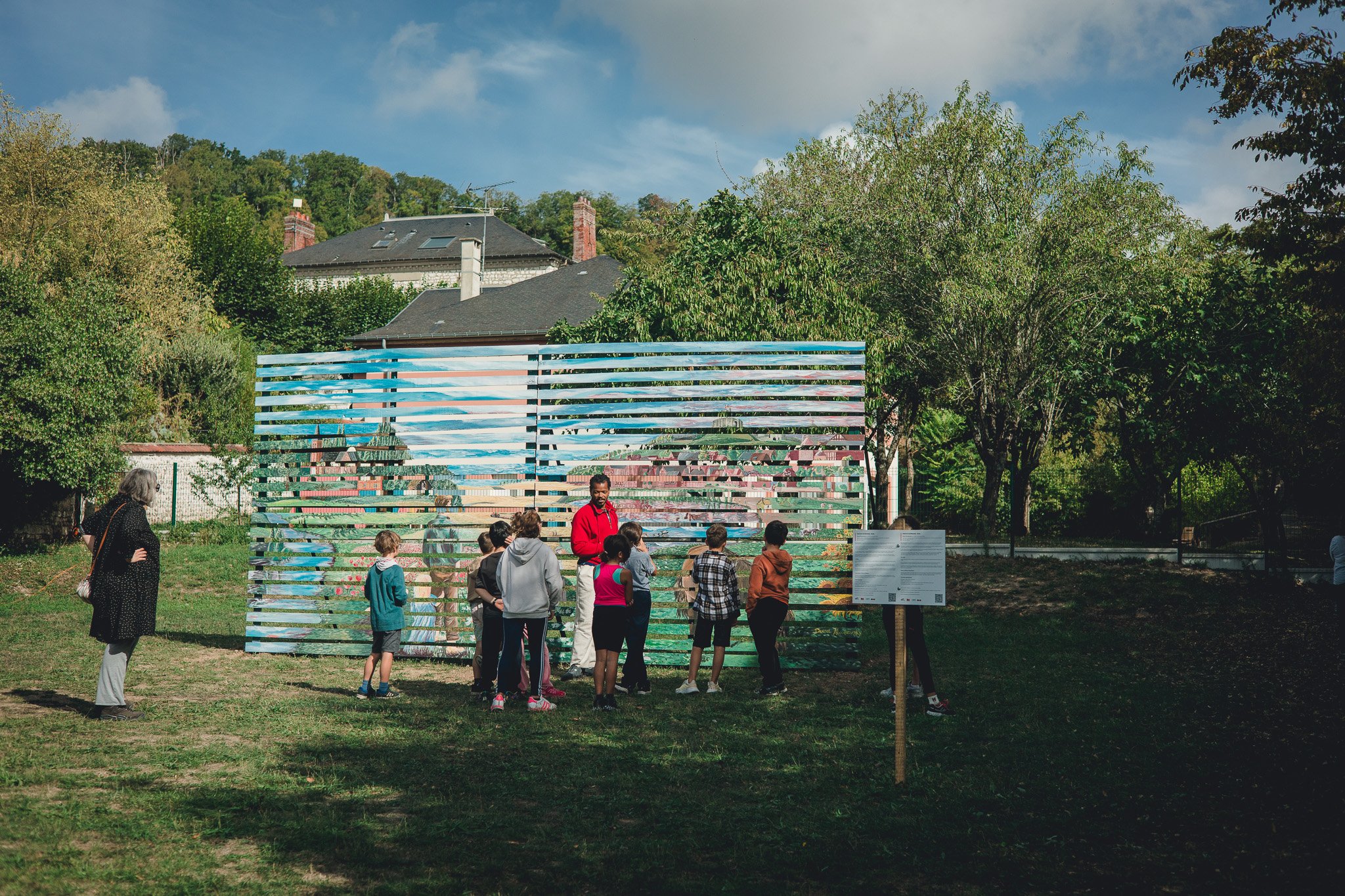 Groupe de personnes, principalement des enfants, regardant une structure d'art public colorée dans un parc avec des arbres et des maisons en arrière-plan.