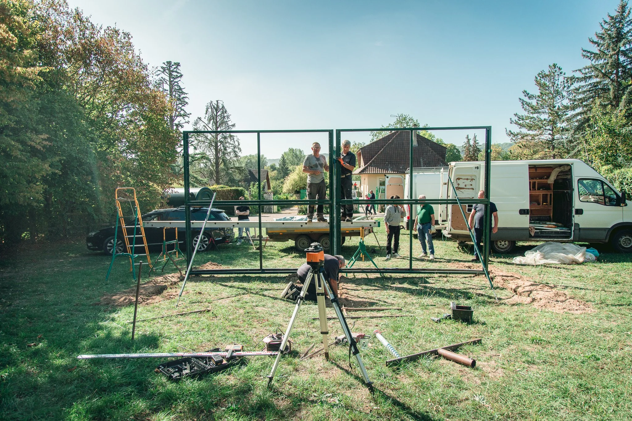 Groupe de personnes construisant une structure métallique en plein air, avec un camion blanc, des outils, et des arbres en arrière-plan.