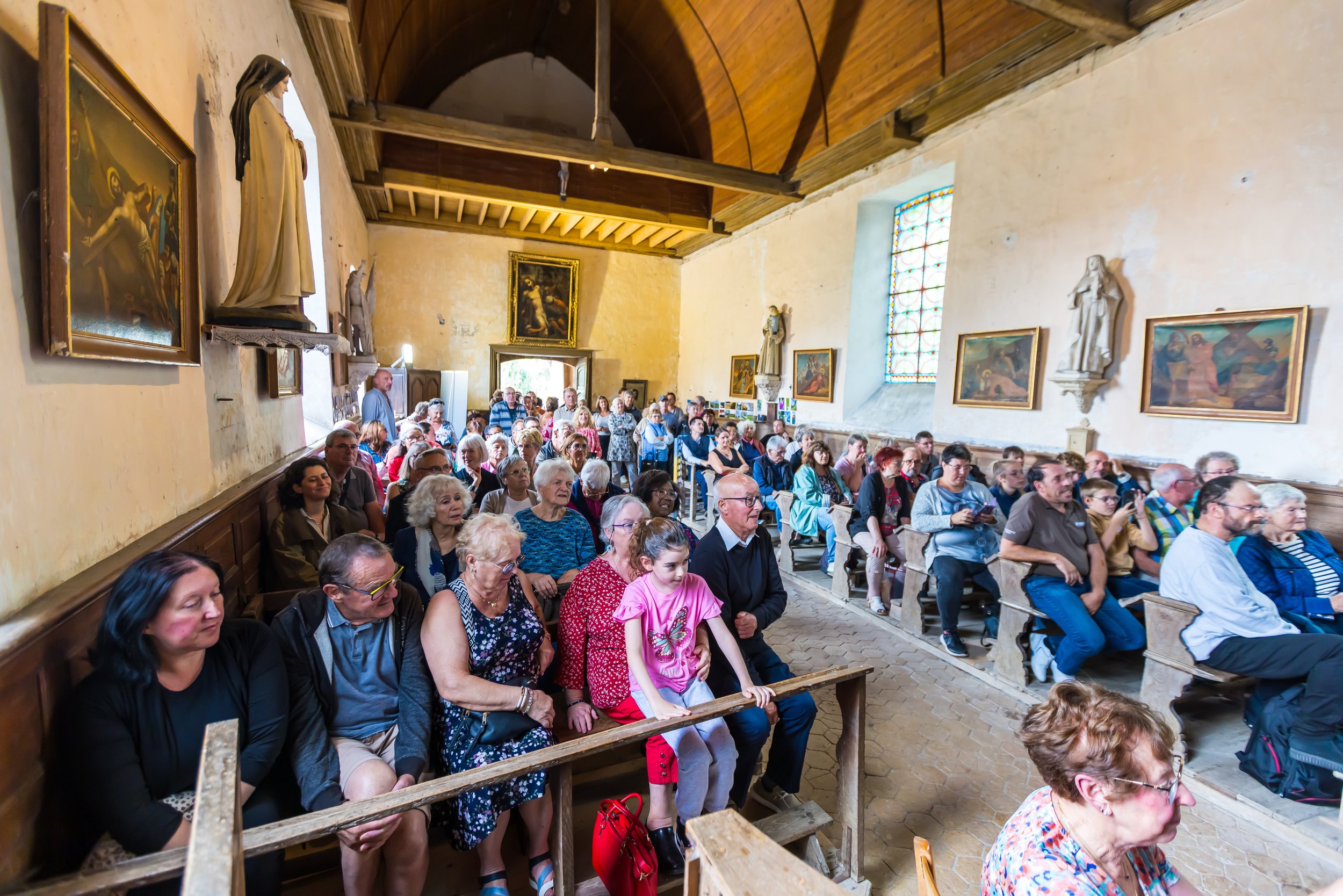 Une salle d'église remplie de personnes assises, avec des statues religieuses et des peintures accrochées aux murs, et des vitraux colorés.