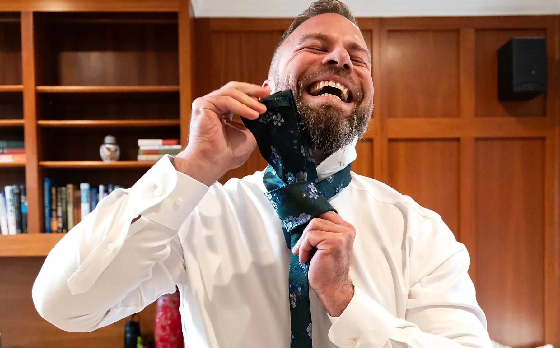 Groom smiling and laughing while putting on a blue floral necktie in a room with wooden bookshelves.