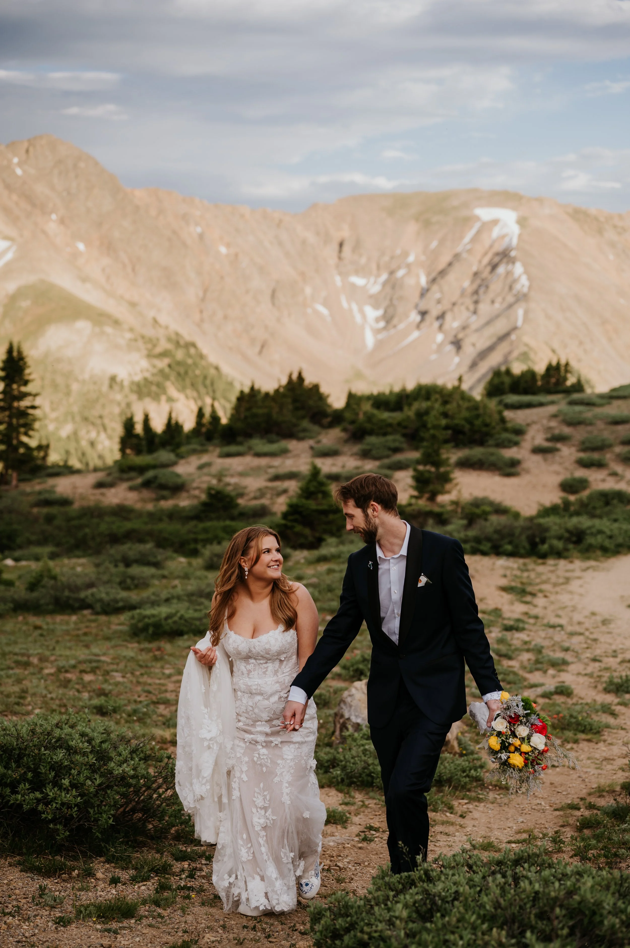 A bride and groom walking hand in hand in a mountainous outdoor setting, the bride in a lace wedding gown holding a bouquet, and the groom in a black suit and white shirt, smiling at each other. Colorado wedding photographer, loveland pass