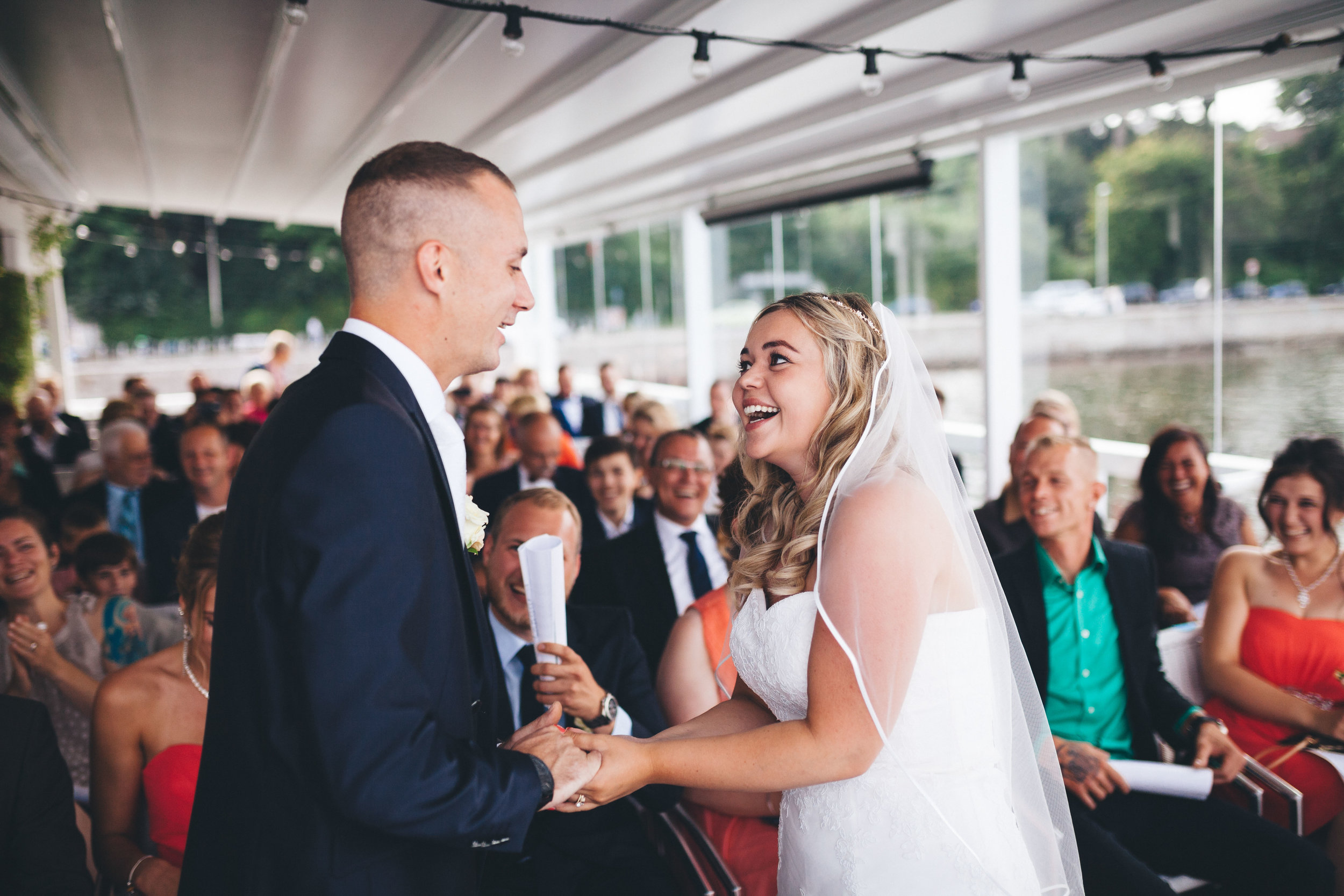 Braut und Bräutigam halten sich bei einer Hochzeit die Hände, schauen sich an und lachen. Sie sind energisch. Im Hintergrund sieht man die Hochzeitsgäste in der Seebar Kiel.