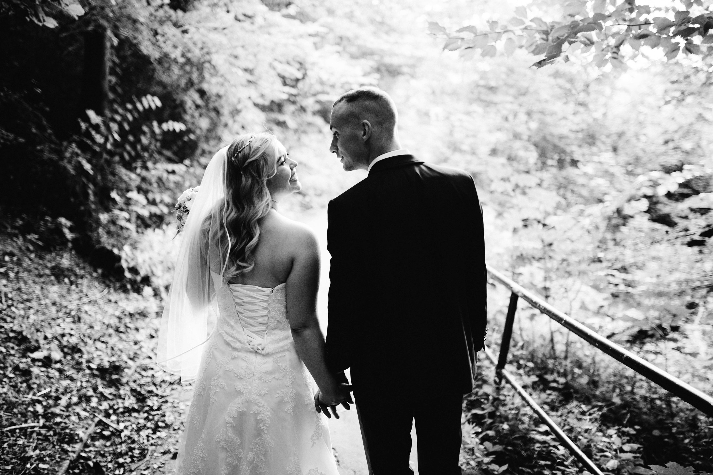 Ein Hochzeitspaar läuft über einen Wald weg. Sie halten Händchen und schauen sich an. Das Foto ist Schwarz Weiß. Die Aufnahme entstand bei eine Fotoshooting in Kiel