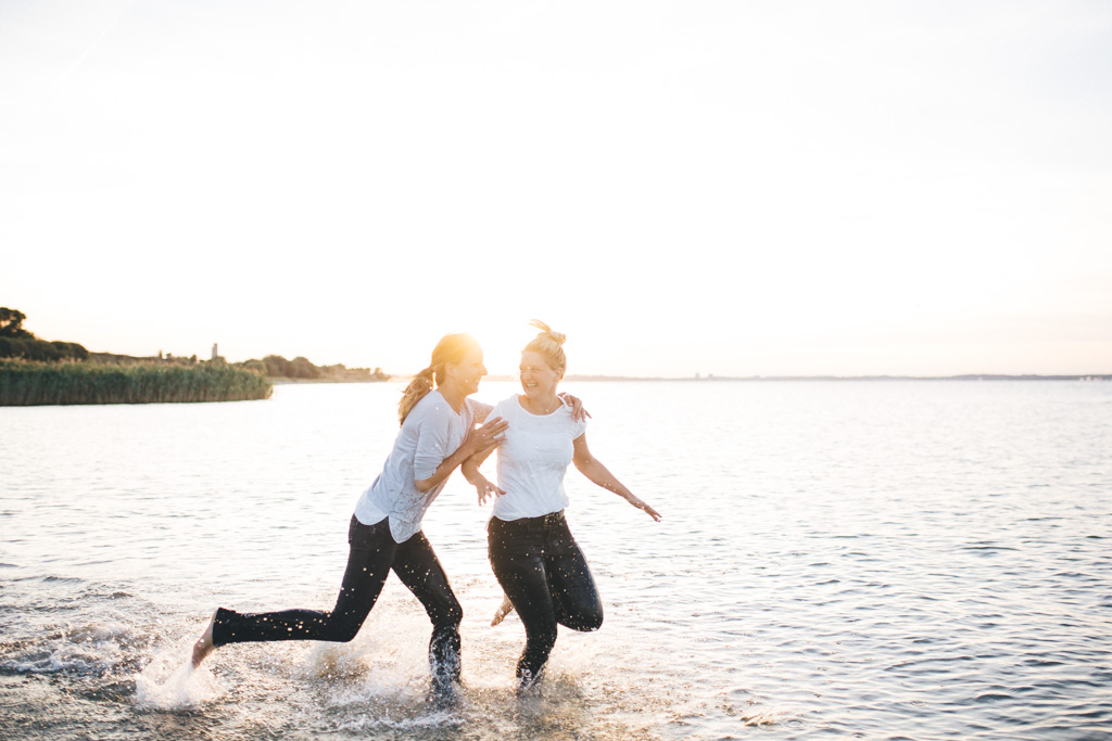 zwei freundinen laufen am Strand im Ortsteil Stein von Kiel, durchs Wasser. Im Hintergrund geht die Sonne unter. Das Foto entstand bei einem Freundinen Fotoshooting in Kiel von Fotograf Phil Schreyer.