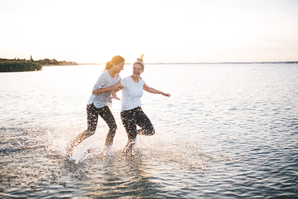 zwei freundinen laufen am Strand im Ortsteil Stein von Kiel, durchs Wasser. Im Hintergrund geht die Sonne unter. Das Foto entstand bei einem Freundinen Fotoshooting in Kiel von Fotograf Phil Schreyer.