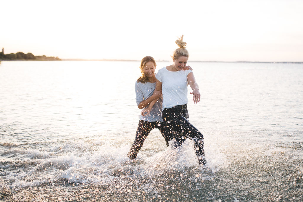 zwei freundinen laufen am Strand im Ortsteil Stein von Kiel, durchs Wasser. Im Hintergrund geht die Sonne unter. Das Foto entstand bei einem Freundinen Fotoshooting in Kiel von Fotograf Phil Schreyer.