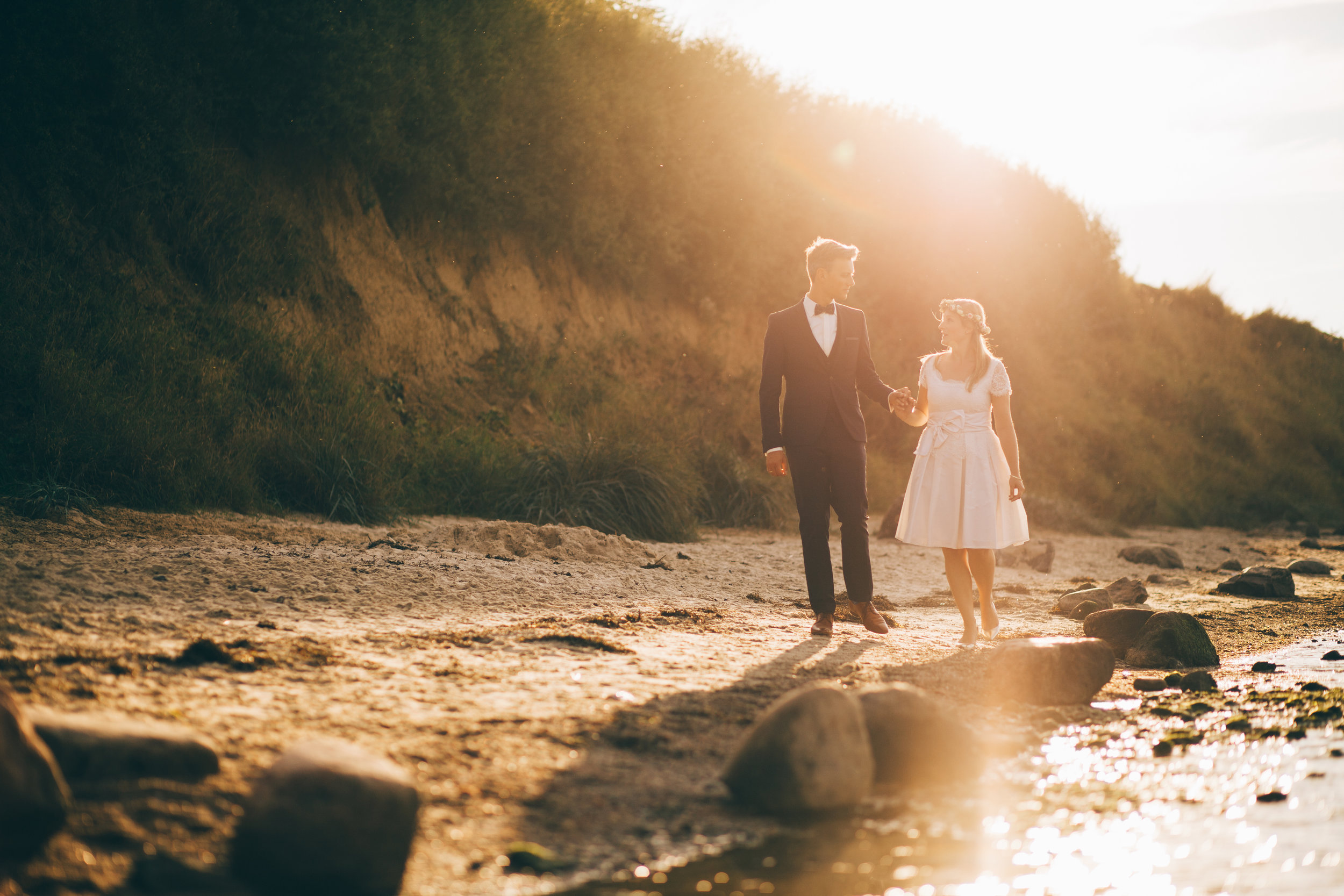 Hochzeitspaar an der Steilküste in Stein bei Kiel. Die Sonne geht unter und sie laufen Hand in Hand am Wasser entlang. Foto von Fotograf Phil Schreyer