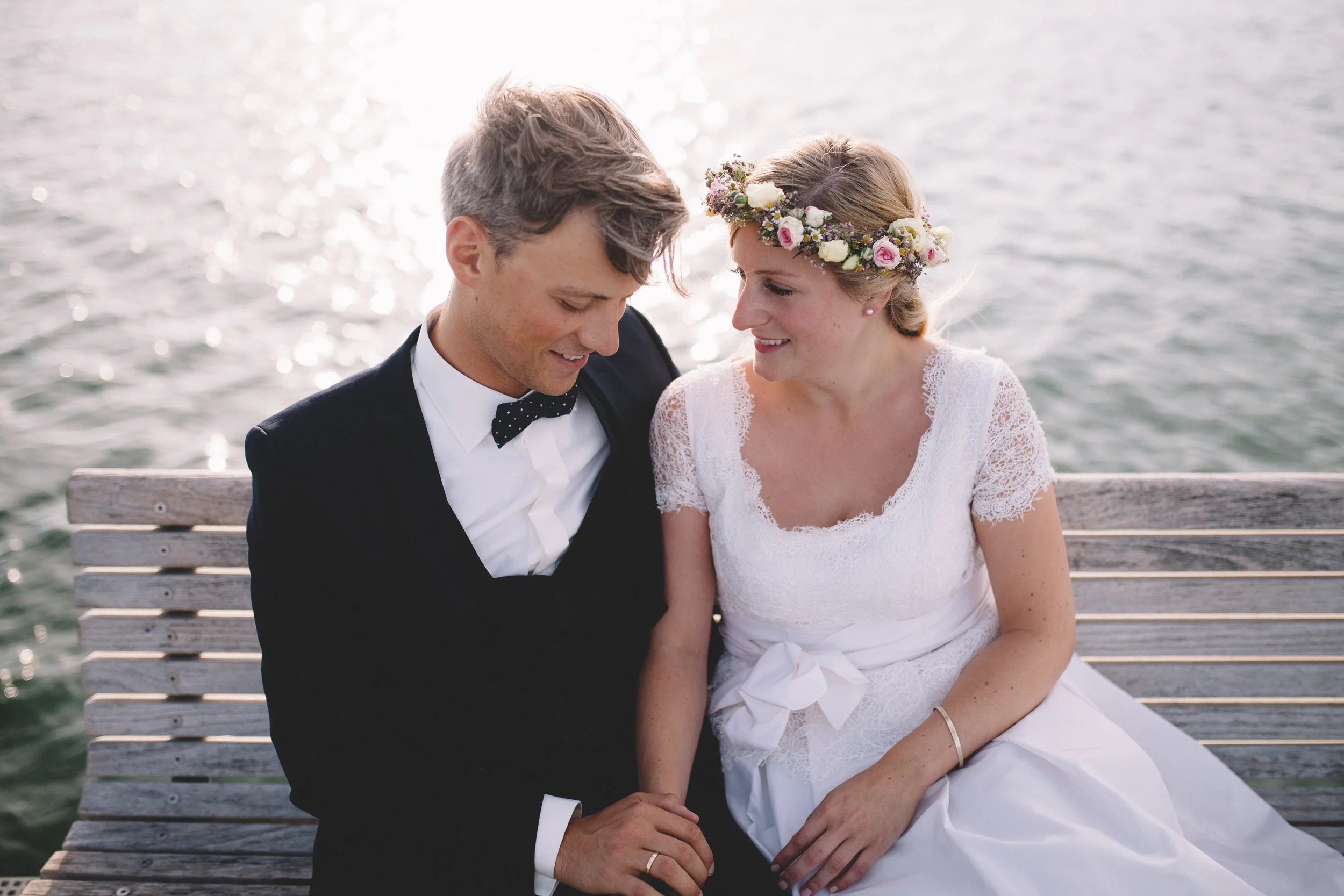 Hochzeitspaar sitzt auf einer Bank am Wasser und schaut sich an. Beide lachen und die Braut hat einen Blumenkranz im Haar. Das Foto wurde von Fotograf Phil Schreyer geschossen