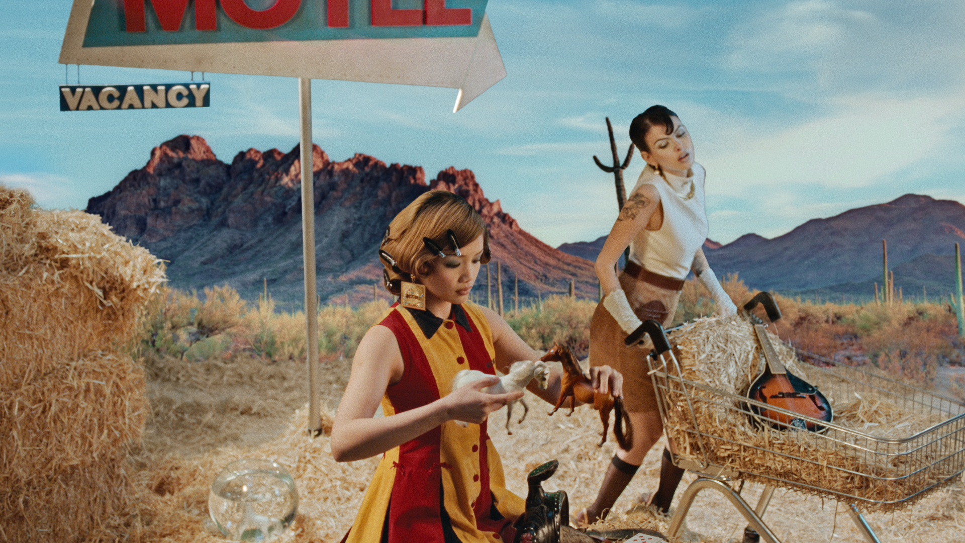 Fashion campaign for Moschino featuring two models in a stylised desert motel scene with hay bales, vintage props and a mountain backdrop, photographed by cinematographer Louis Brennan.