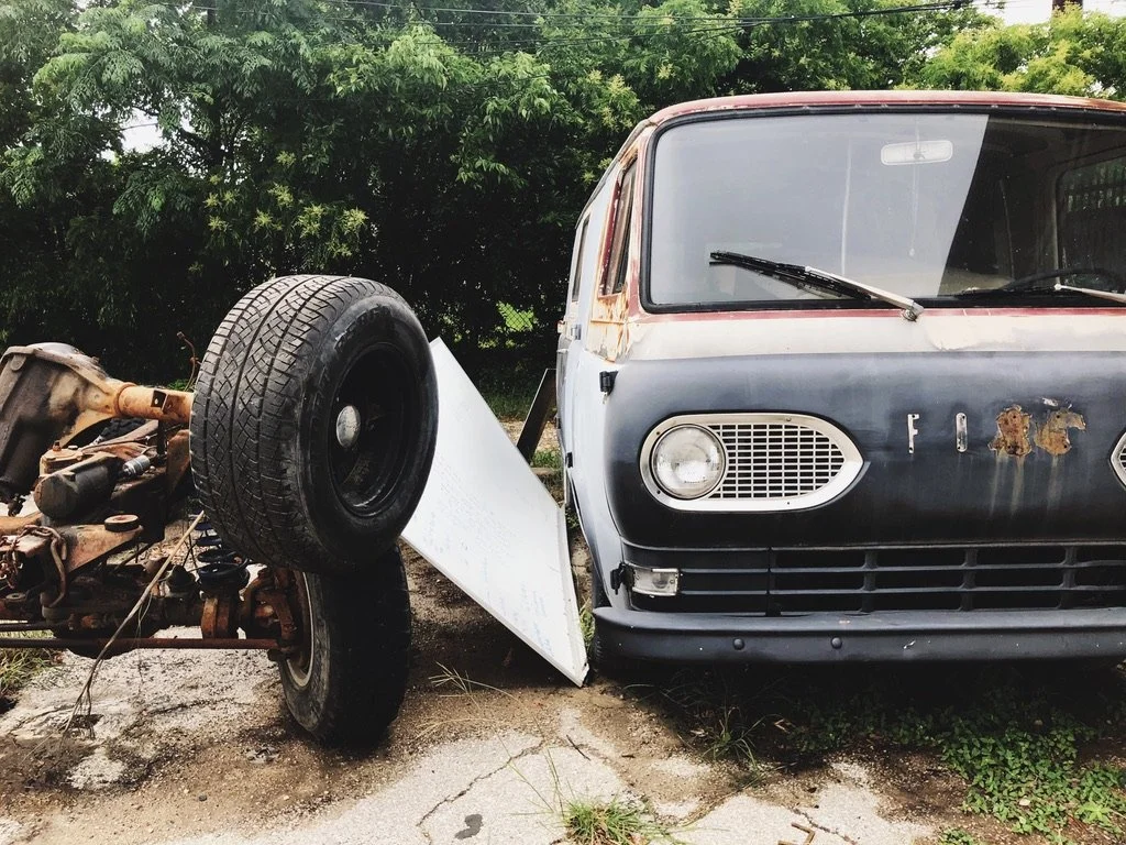An old, rusty van with peeling paint parked next to a disassembled car chassis with a spare tire on top, outdoors near green trees.