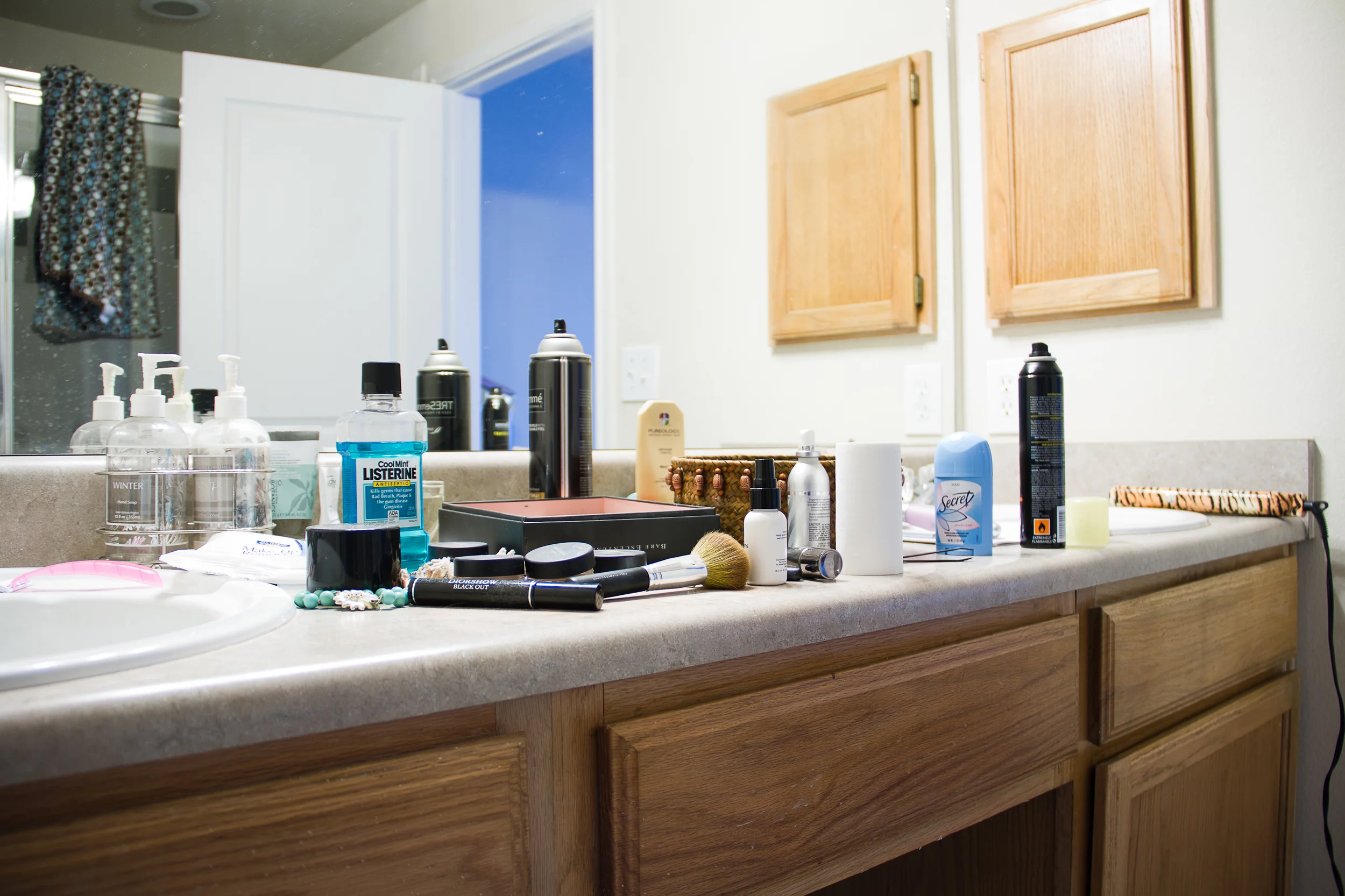 Bathroom countertop cluttered with personal care items, including a bottle of Listerine mouthwash, hair spray cans, a makeup brush, and various toiletries, with a mirror and wooden cabinets in the background.