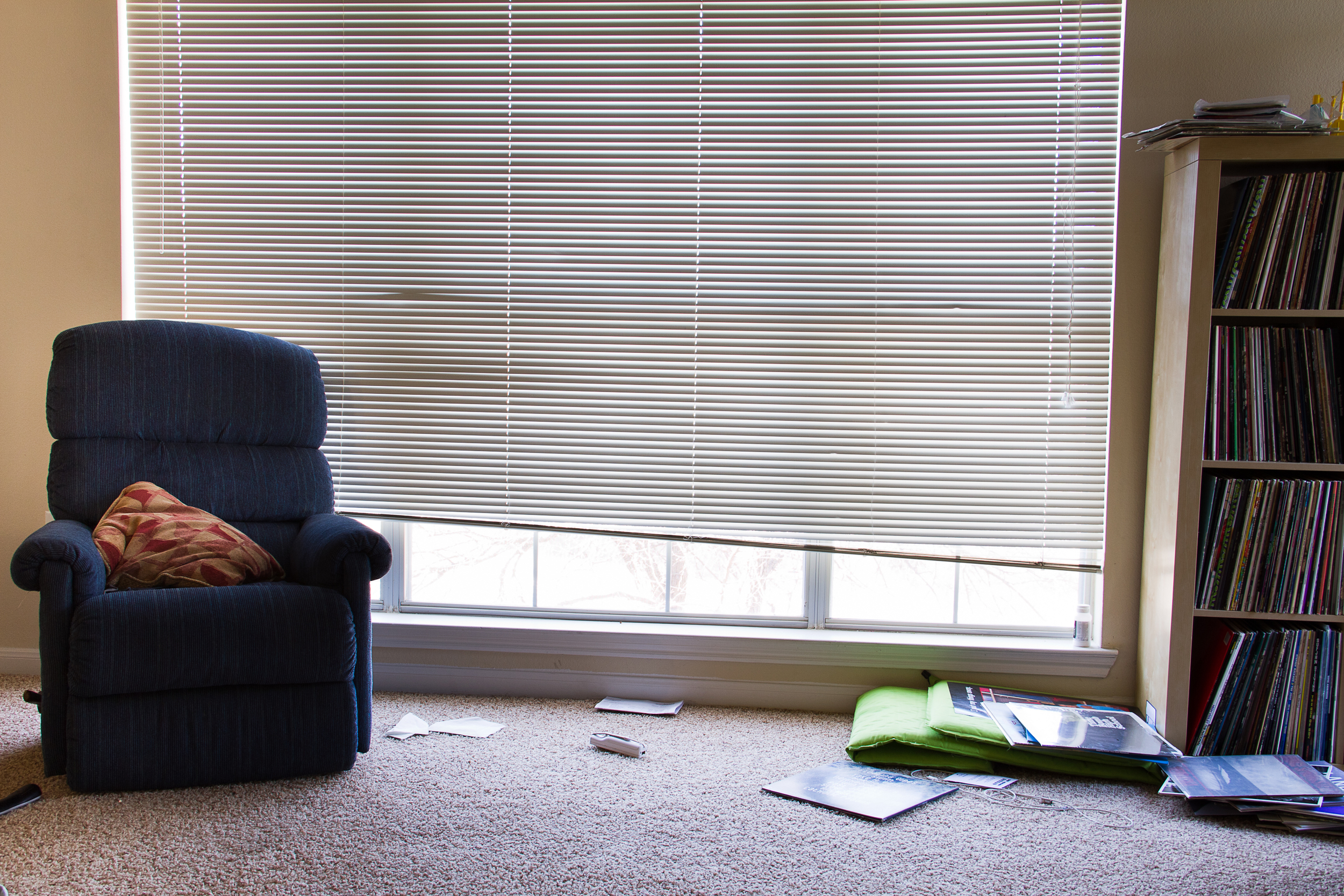 Living room with a navy armchair, a window with closed blinds, a bookshelf on the right, and scattered magazines and papers on beige carpet.