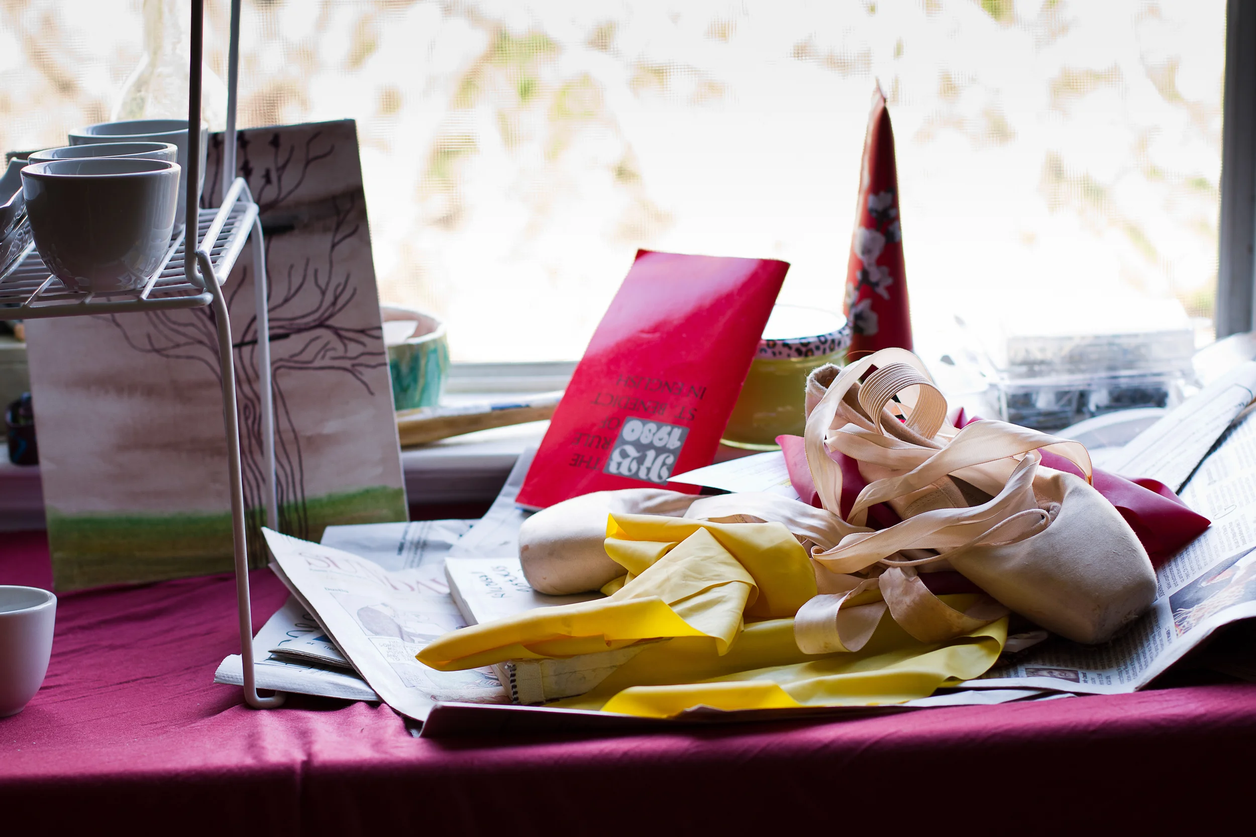 Disorganized table with newspapers, ribbons, a tote bag, a red booklet, party hats, and cups near a window.