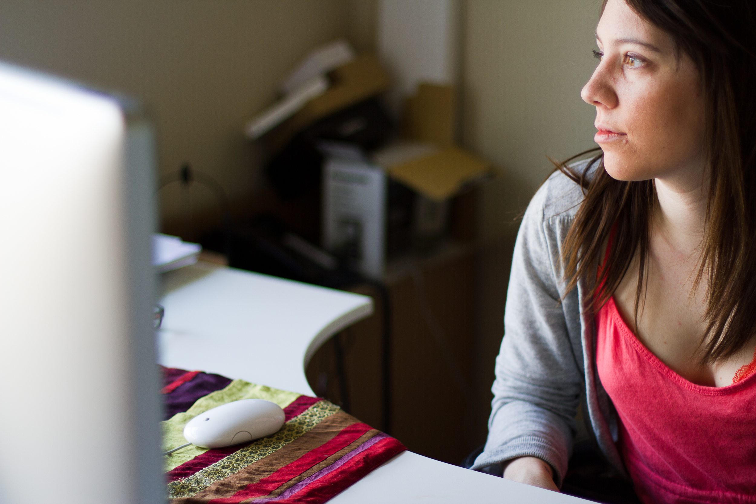 A woman sitting at a desk in front of a computer, looking intently at the screen with natural light illuminating her face. A computer mouse is on a colorful cloth on the desk.