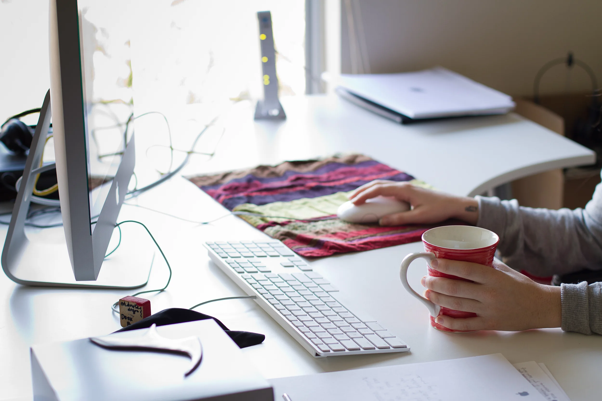 Person using a computer mouse with a coffee mug in hand, at a cluttered office desk with a keyboard, paper, folders, and a monitor.