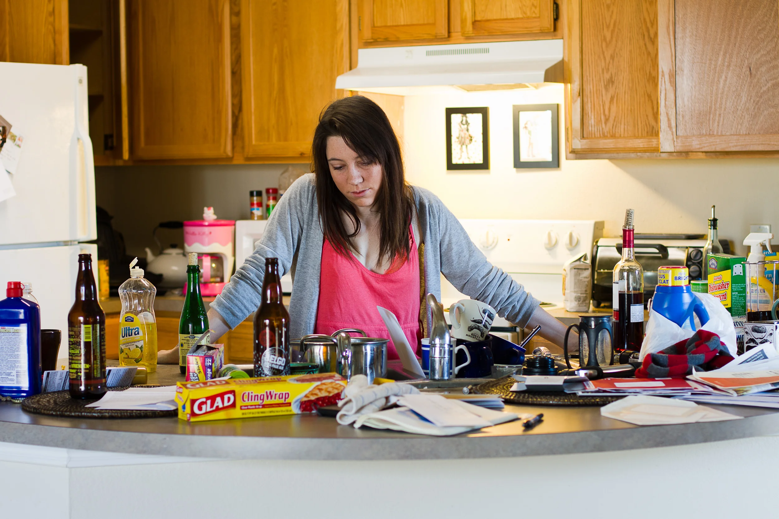 A woman with dark hair wearing a pink shirt and gray hoodie, looking down at a cluttered kitchen counter with various bottles, papers, and kitchen items.