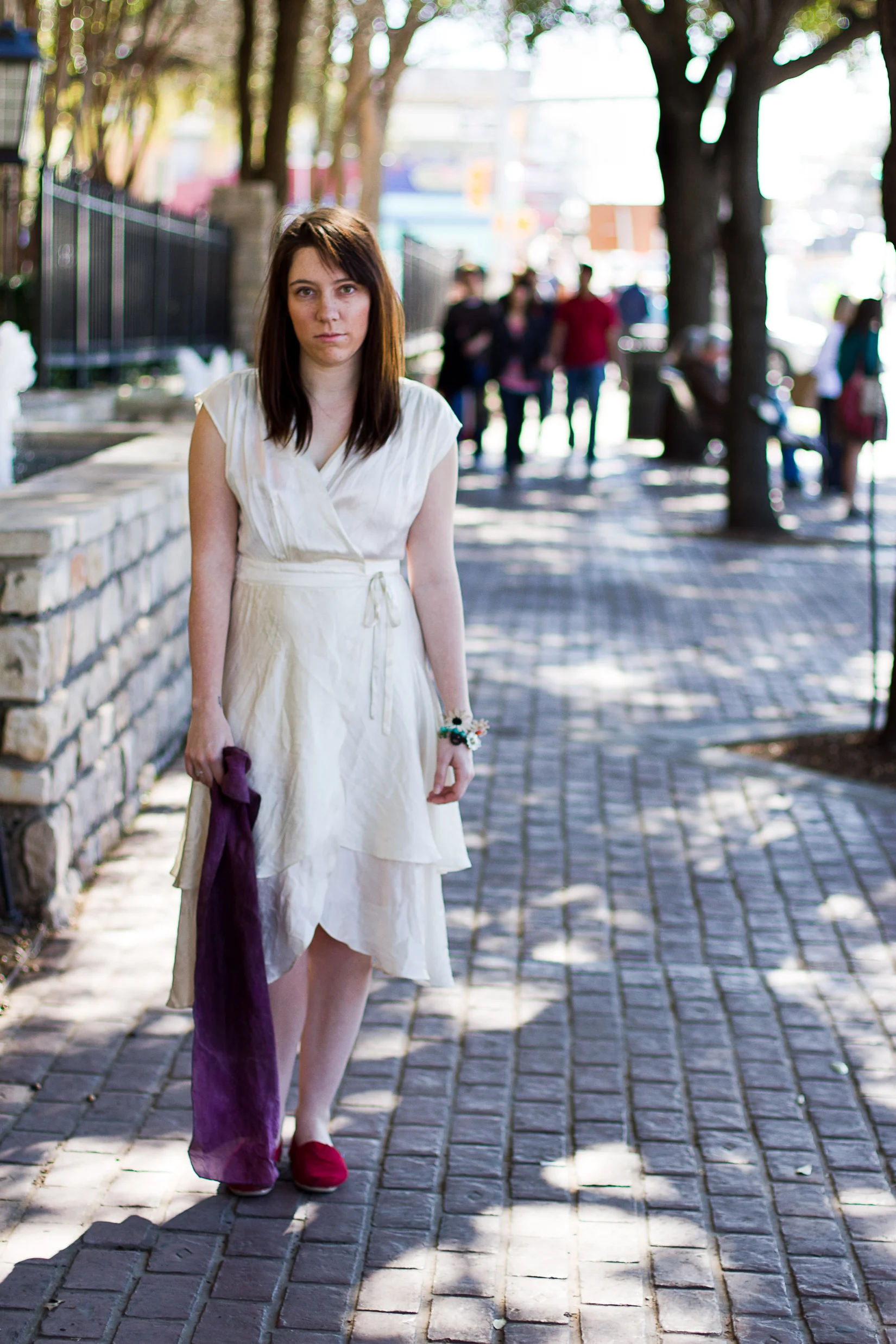 A young woman with dark brown hair wearing a cream-colored dress, holding a purple cloth, walking on a brick sidewalk in an urban setting with trees and a group of people in the background.