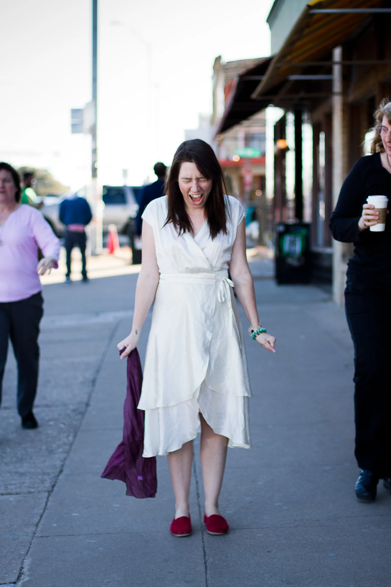 A woman in a white dress and red slippers walking on sidewalk with an expression of excitement or joy, surrounded by other people on the street.