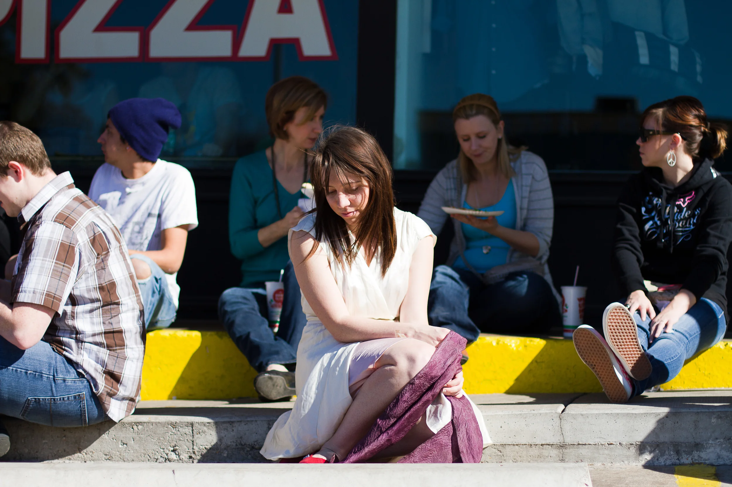 A group of people sitting and standing outside a restaurant, with one woman in a white dress sitting on a curb in the foreground, holding and adjusting her purple jacket.