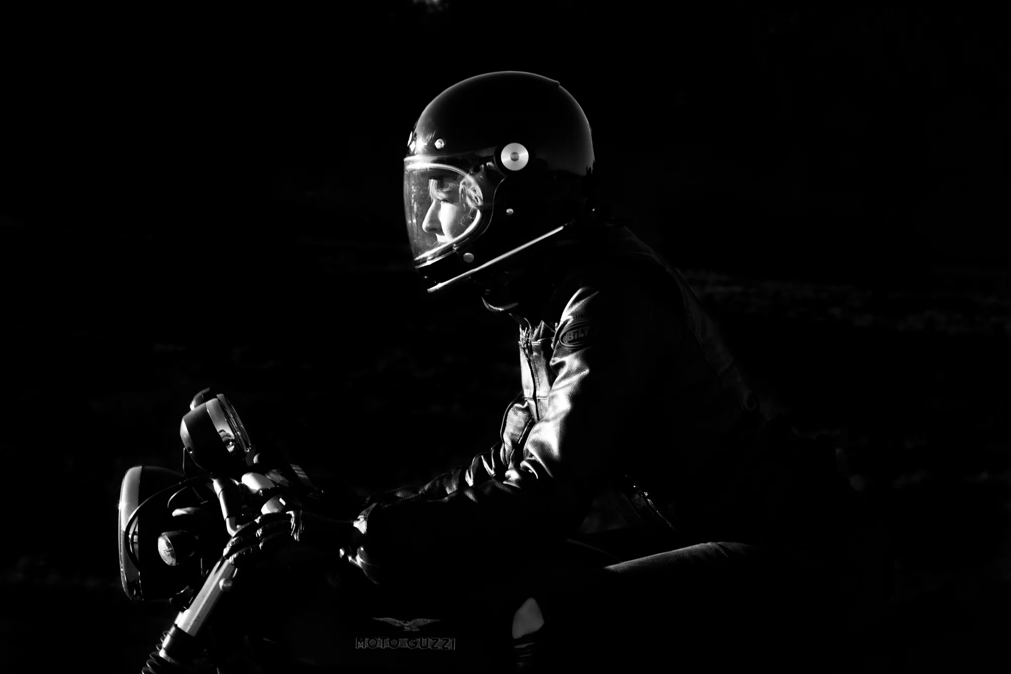 A person wearing a helmet and leather jacket riding a motorcycle in a dark setting.