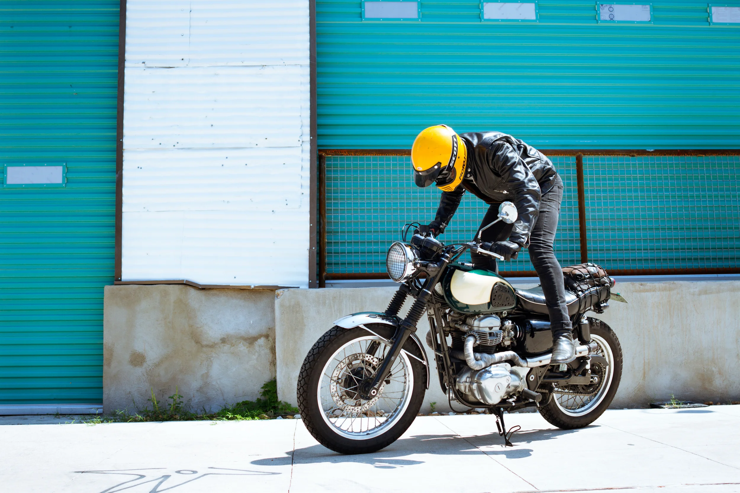 Person in black leather jacket and yellow helmet riding a vintage motorcycle in front of a colorful industrial building with metal siding.