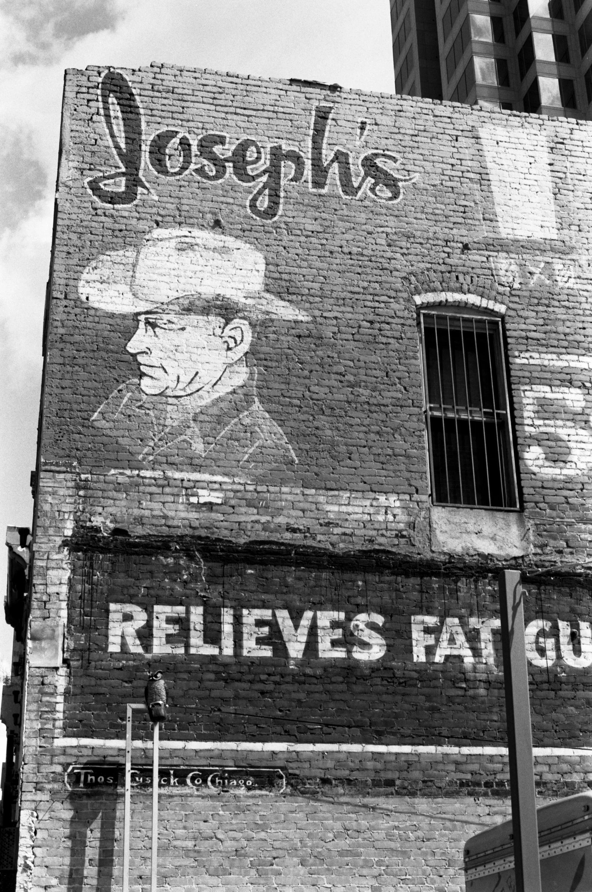 Black and white photo of a brick building with a mural that says 'loose' and a drawing of a man wearing a hat. Part of a street sign and two light poles are visible in front of the building.
