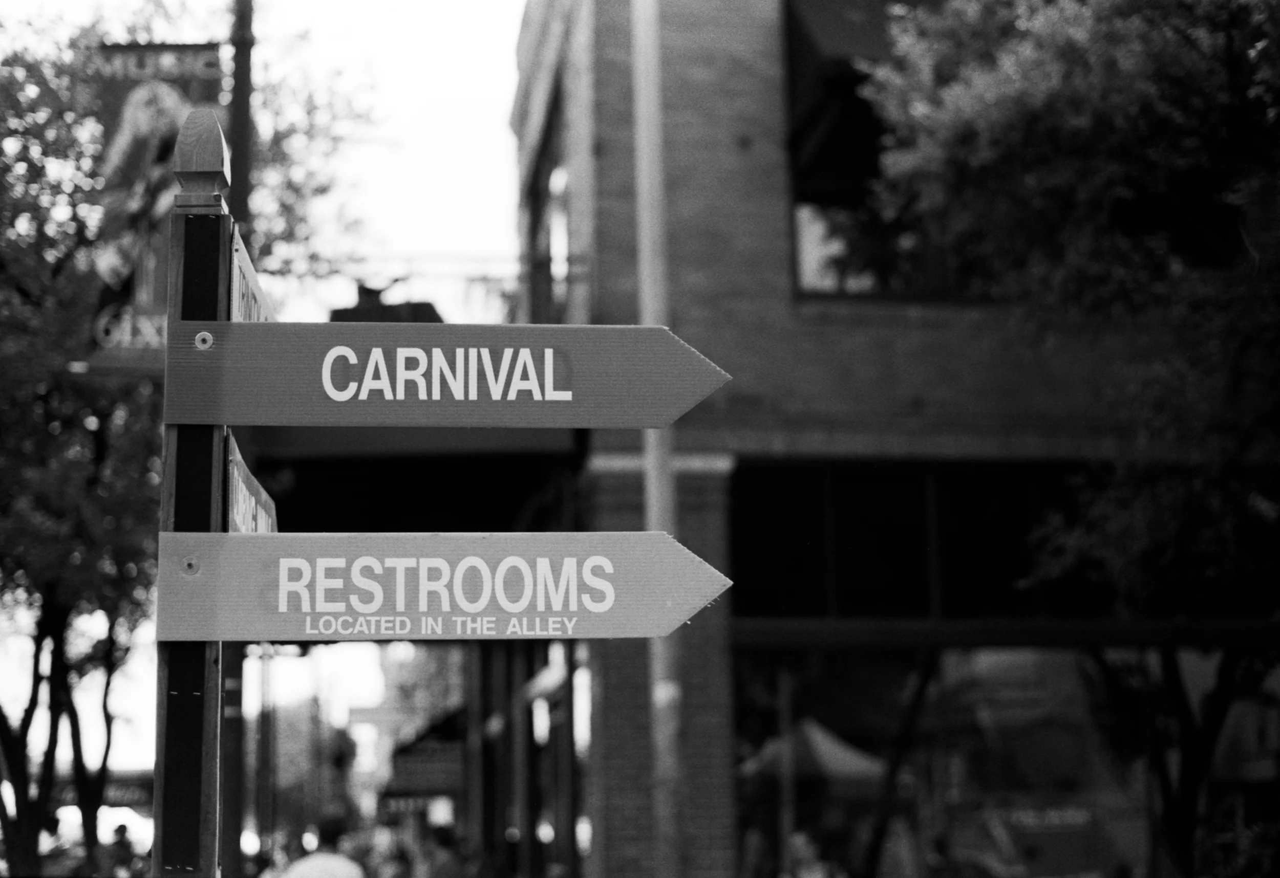 Arrow-shaped sign pointing to the carnival, with another sign below indicating restrooms located in the alley, all in black and white.