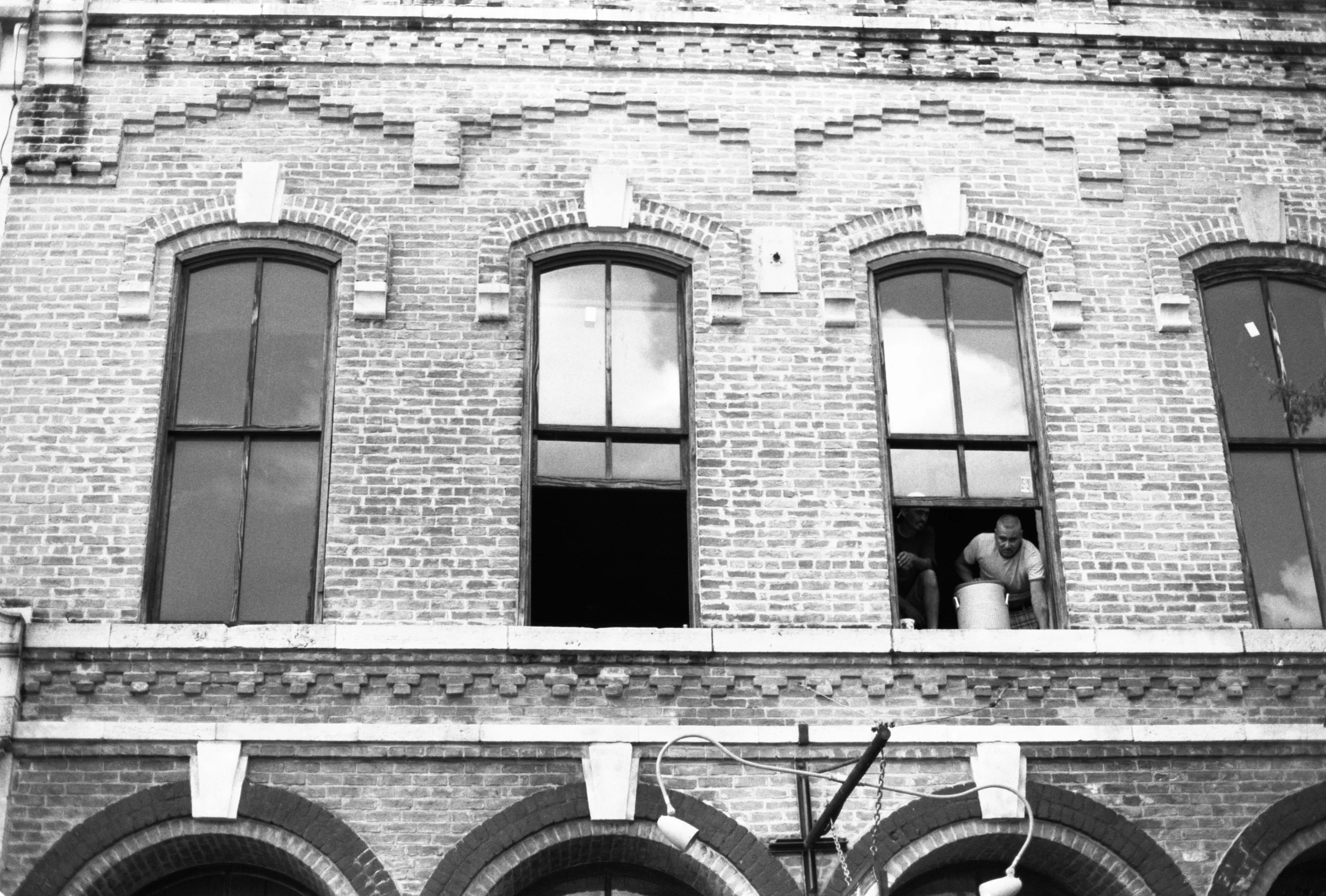 Black and white photo of a brick building with three arched windows on the upper floor, with two men looking out of one of the windows.