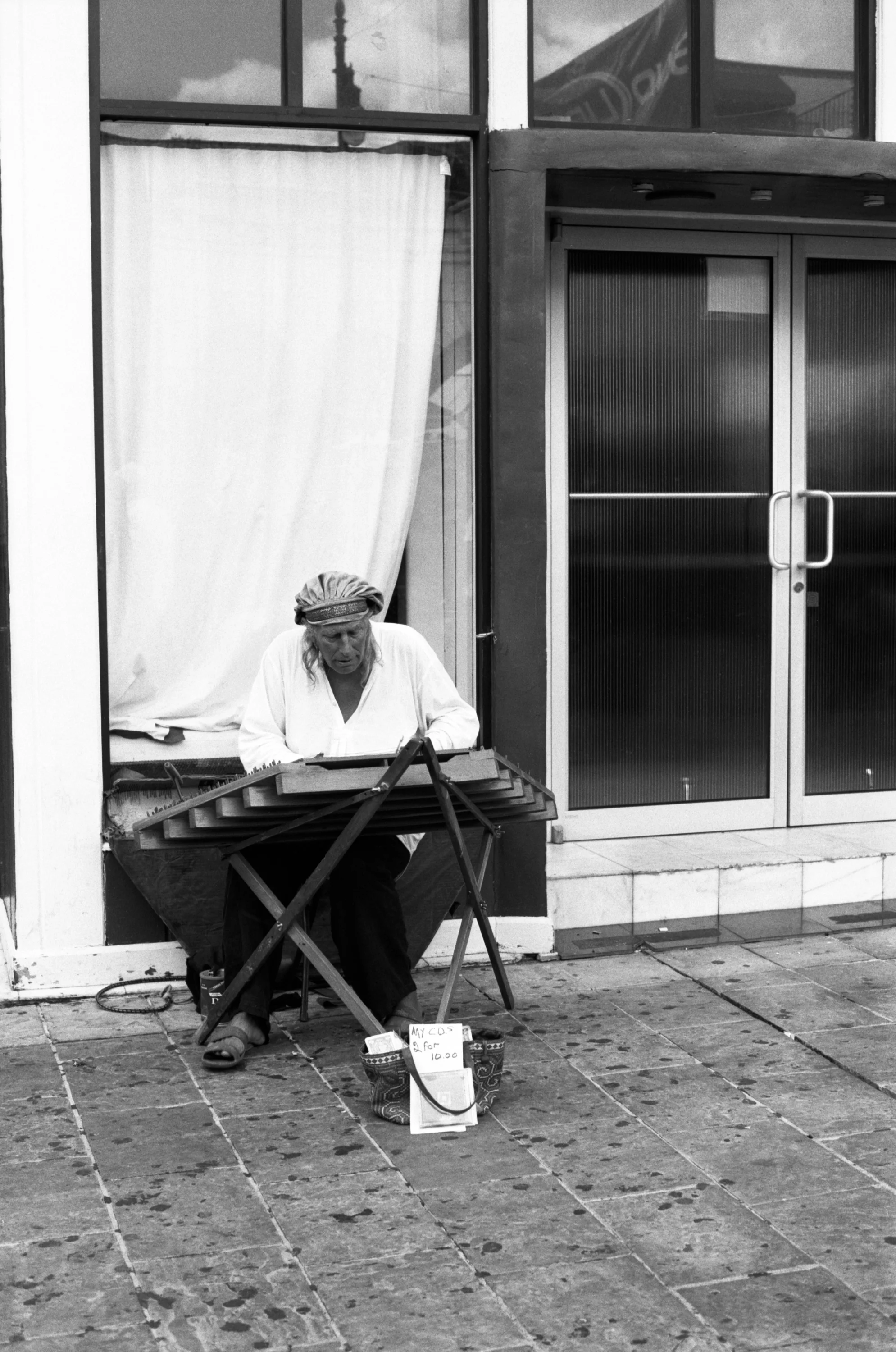 A woman sitting outside a storefront, which has a large glass window and glass door, she appears to be selling handcrafted objects, with a small sign on the ground reading 'My Co's 2 for 1000' and a bucket with more signs or labels.