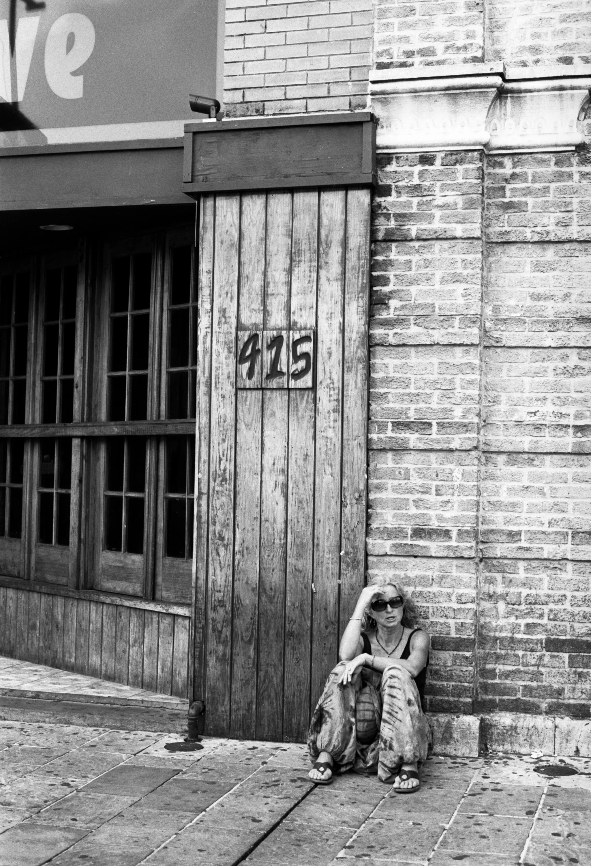 A woman sitting on the sidewalk next to a brick building with a wooden gate, wearing sunglasses, patterned pants, and sandals.