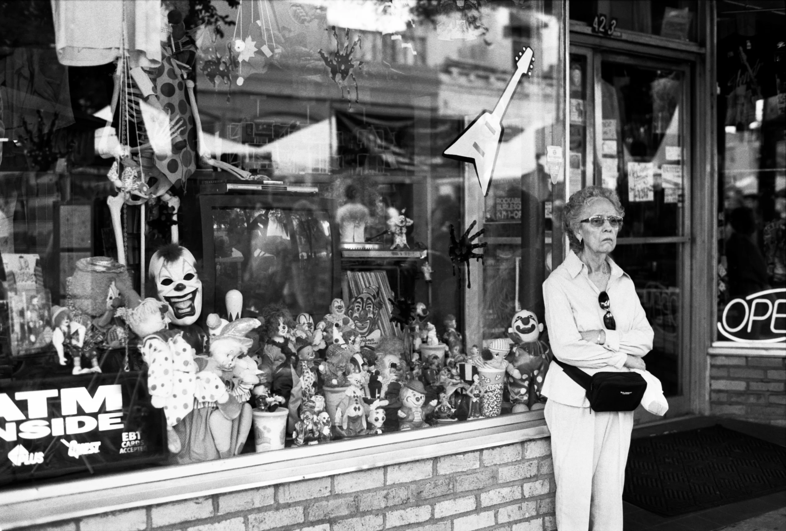 A storefront window displaying various clown and toy decorations, including clown masks, dolls, and plush toys. An elderly woman with glasses and a white shirt is standing outside the store with her arms crossed. The store has a brick exterior and a 