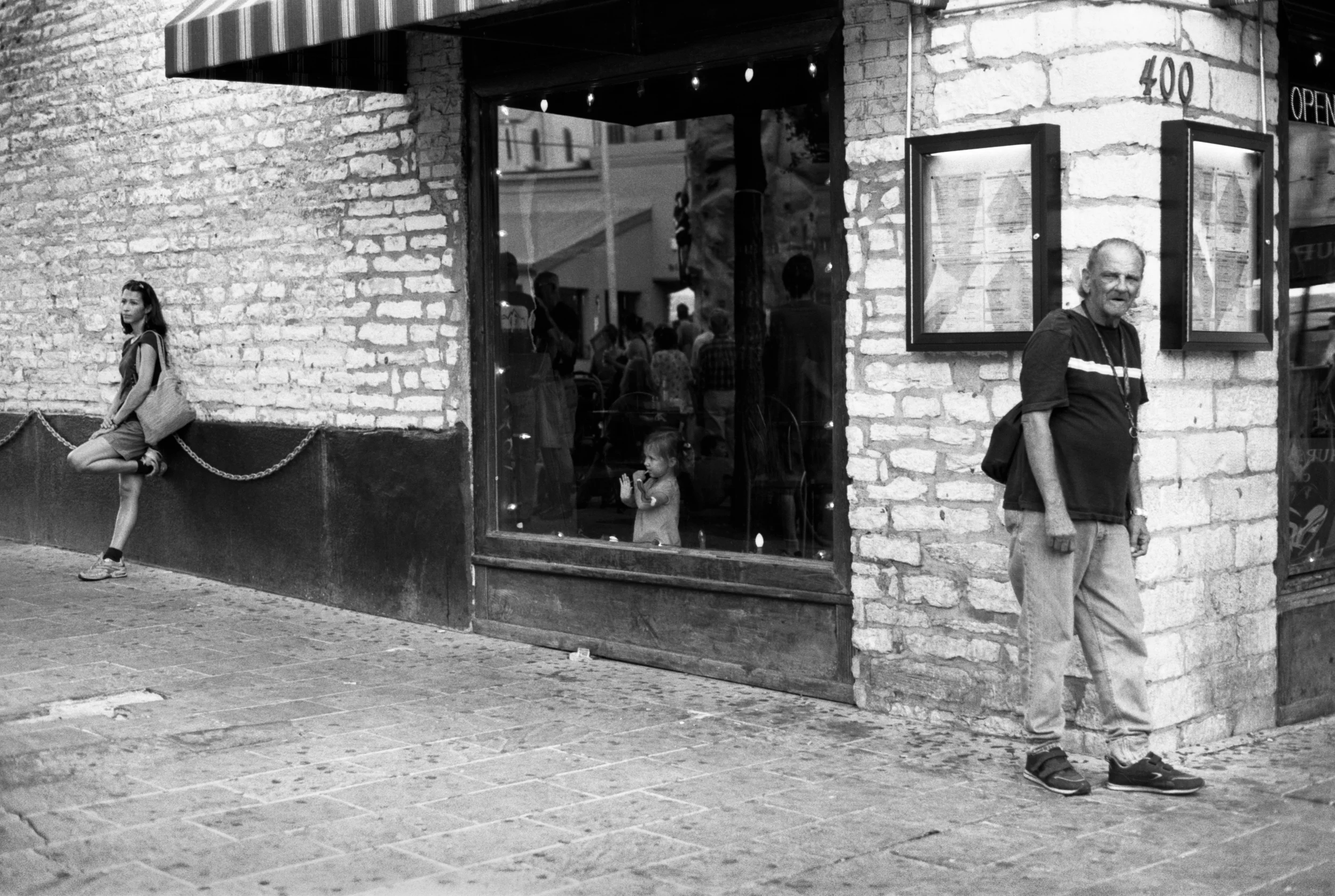 A black and white photo of three people outside a restaurant with a stone wall and large windows. A girl leans against a chain barrier, a young child looks into the window, and an older man stands nearby.