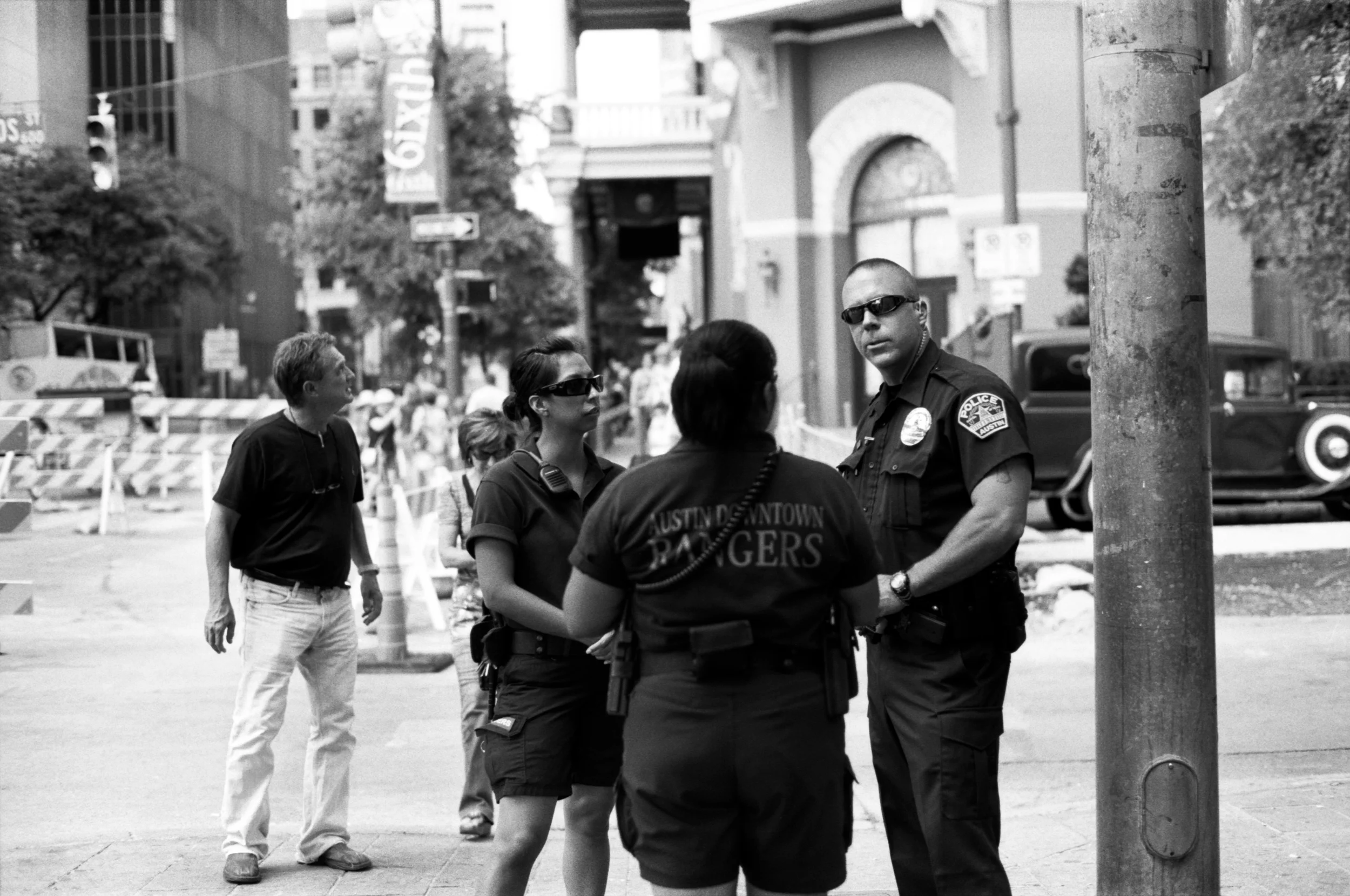 A black and white photo of four police officers, two women and two men, standing and talking on a city street. One male officer with sunglasses is facing the camera. The background shows buildings, trees, and city traffic.
