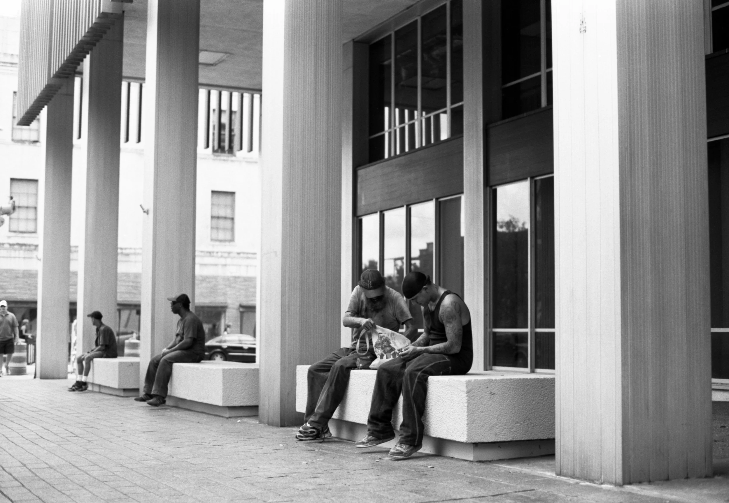 People sitting on benches outside a modern building with vertical metal panels, large windows, and a covered walkway.