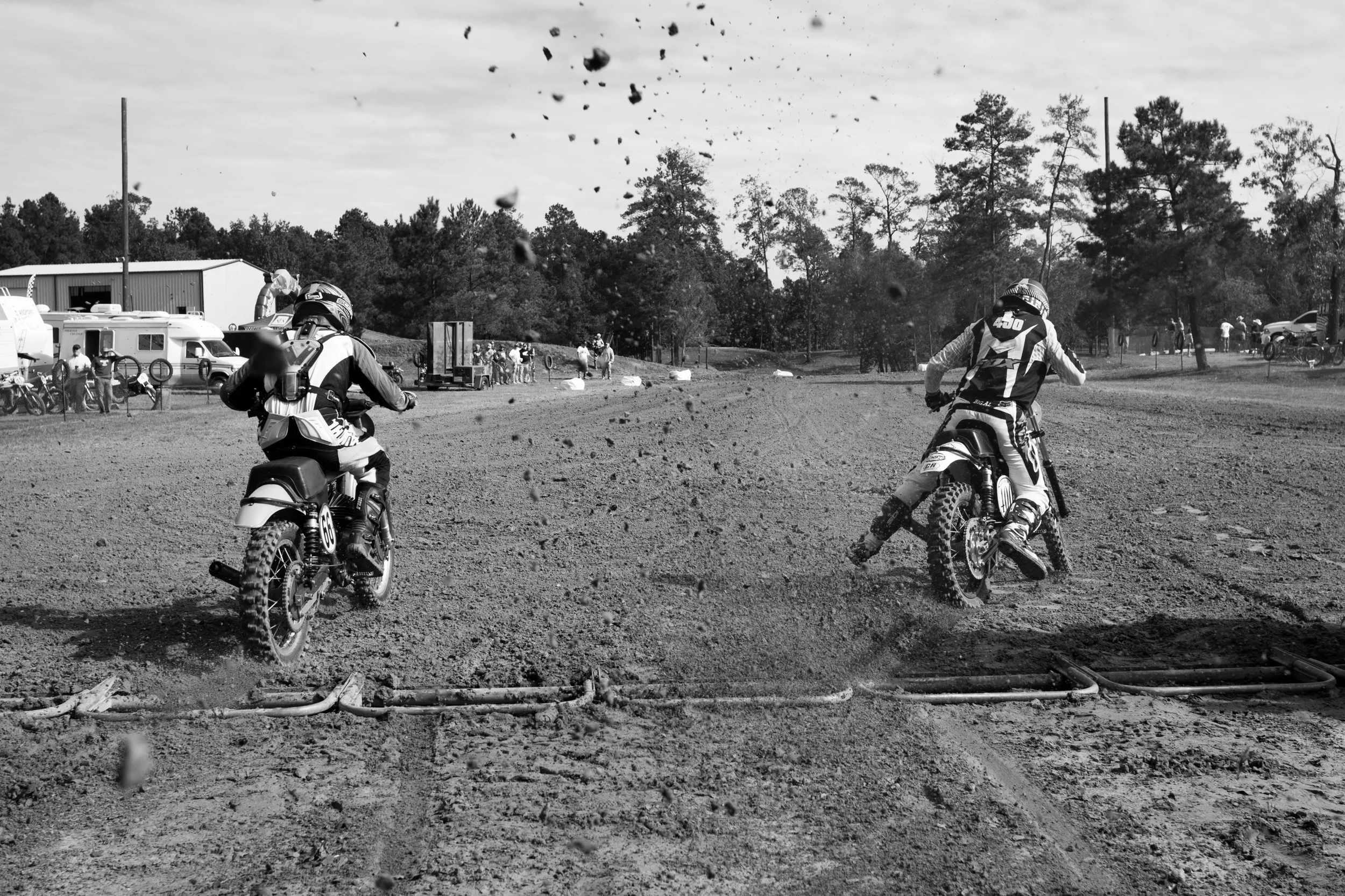 Two dirt bike racers wearing helmets and protective gear on a dirt track, kicking up dirt as they race.
