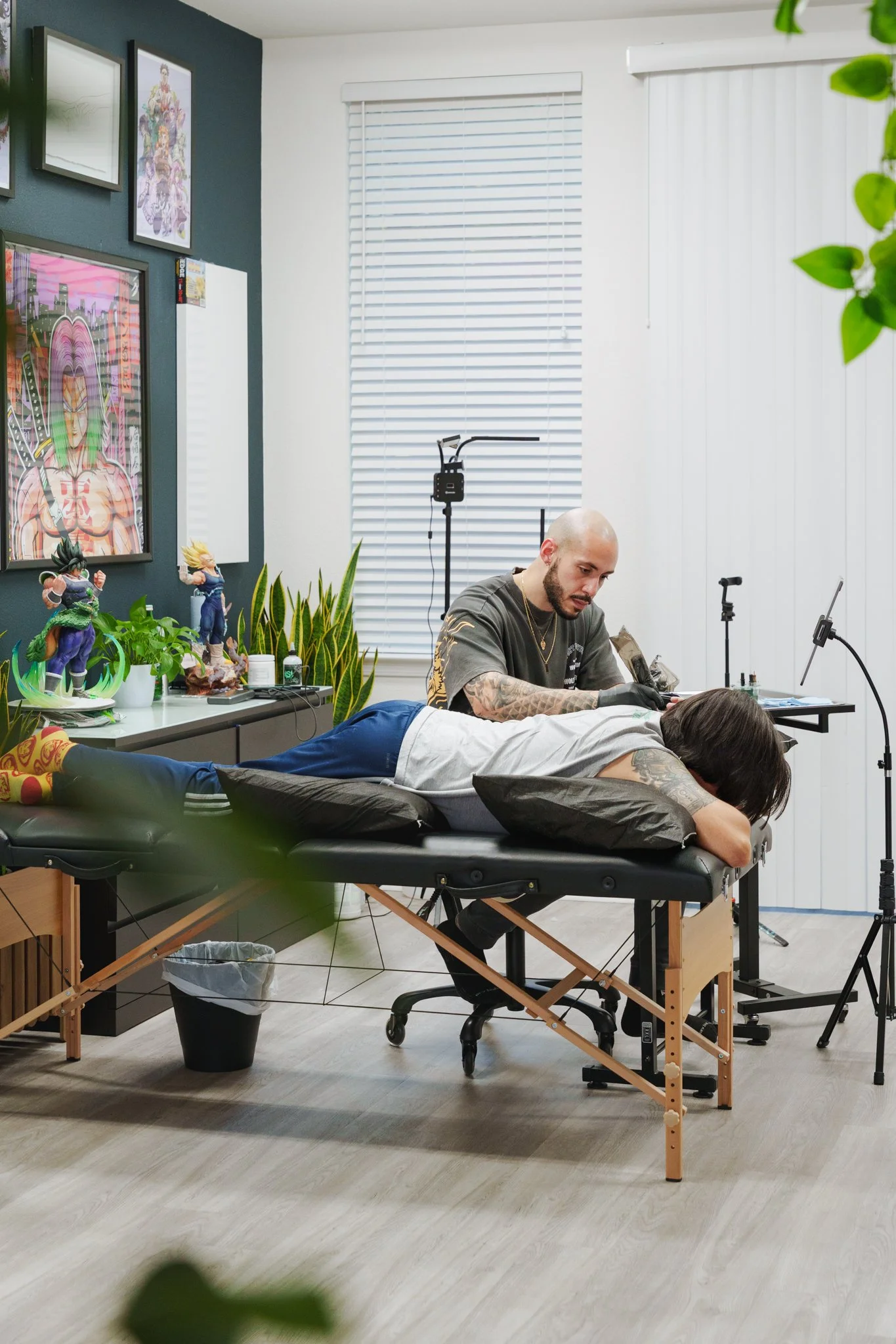 A person with dark hair and tattoos receiving a tattoo on their back while lying face down on a tattoo table. The tattoo artist, a bald man with tattoos on his arms, works on the person. The room has artwork on the walls, a plant, and tattooing equip