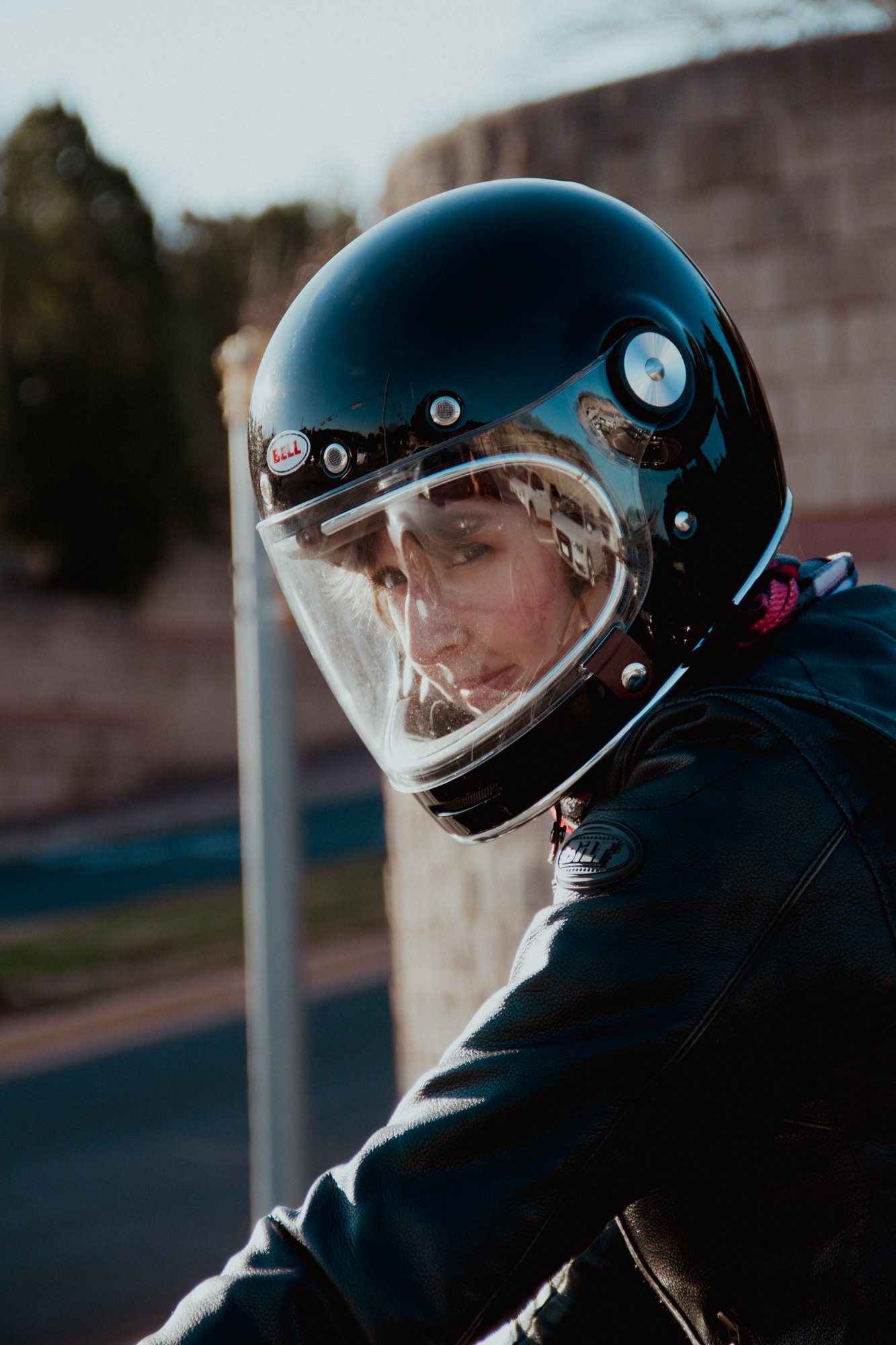 Close-up of a person wearing a black Bell motorcycle helmet with a clear visor, outdoors, with trees and a building in the background.