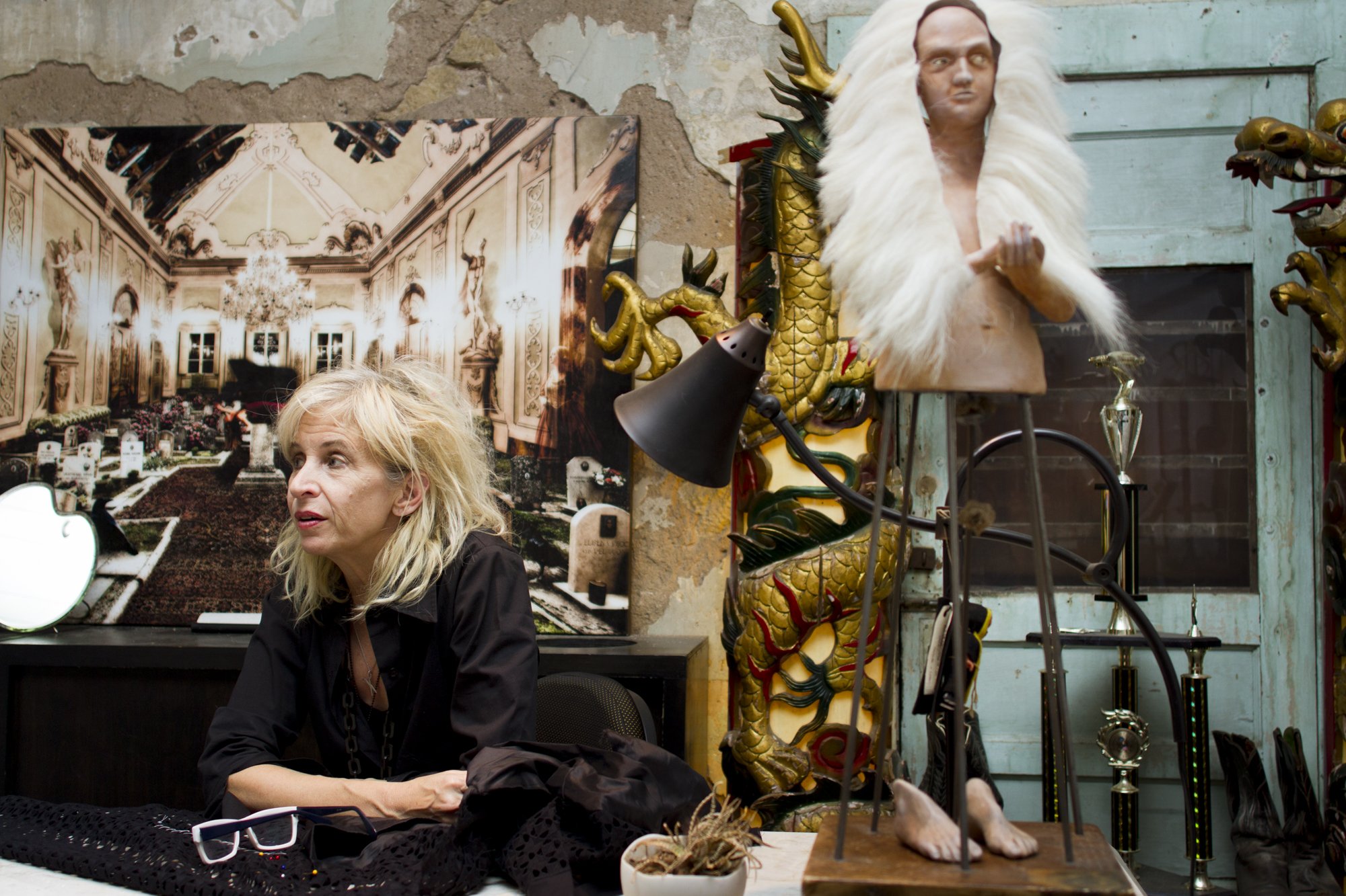 A woman with blonde hair sitting at a table with a black cloth, glasses, and a black shirt. Behind her is a wall with peeling paint and a large ornate photograph of a chandeliered room. On the right, there is a gold and green dragon sculpture with a 
