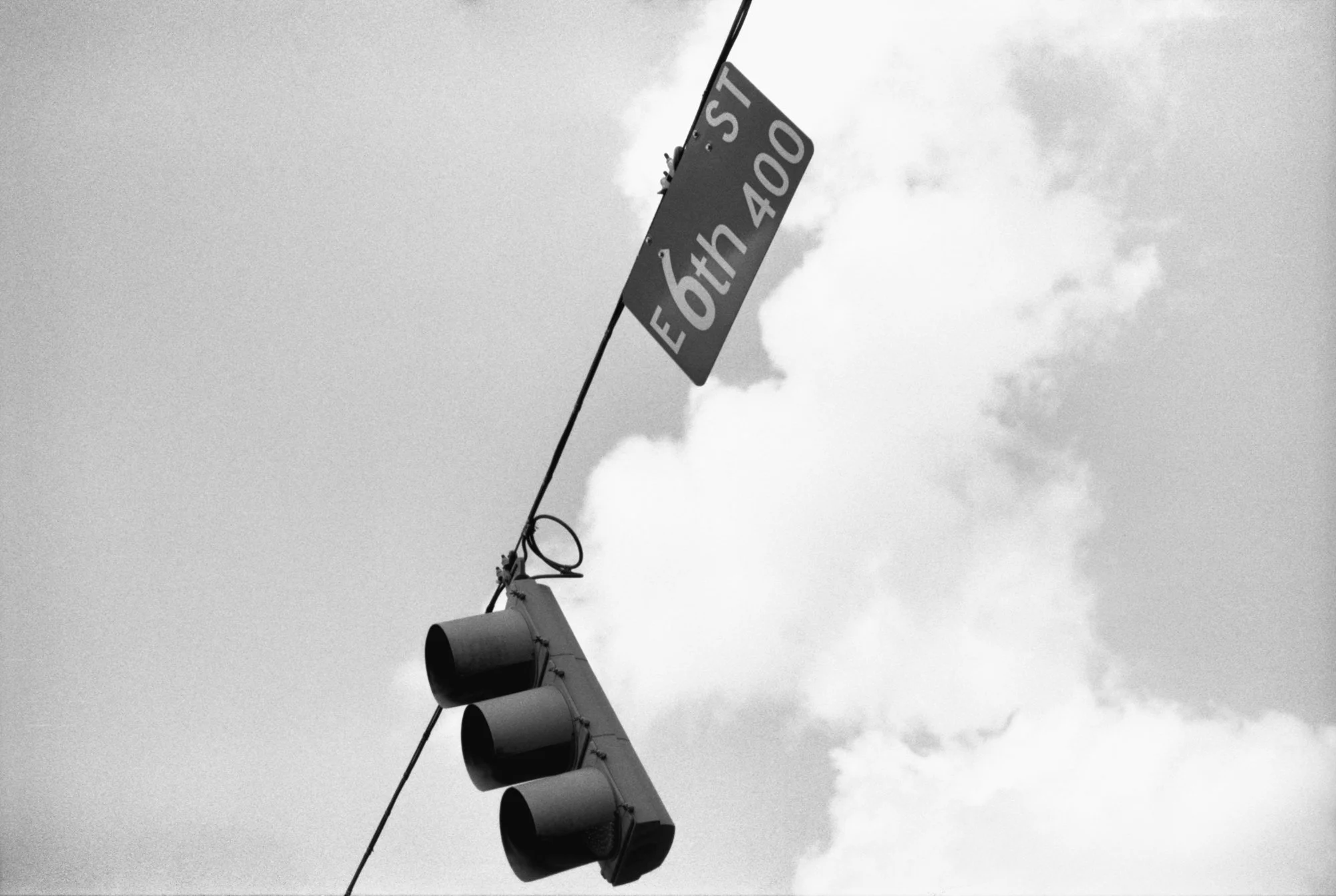 Black and white photograph of a traffic light pole with three traffic lights and a street name sign that reads "E 64th St" against a cloudy sky.