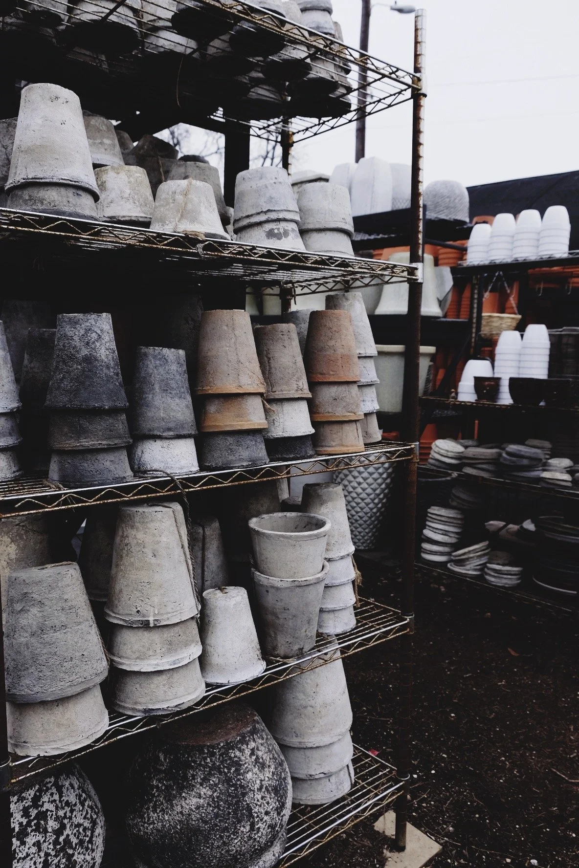 Outdoor shelf displaying weathered flower pots and planters, with additional stacks of pots and dishes nearby.