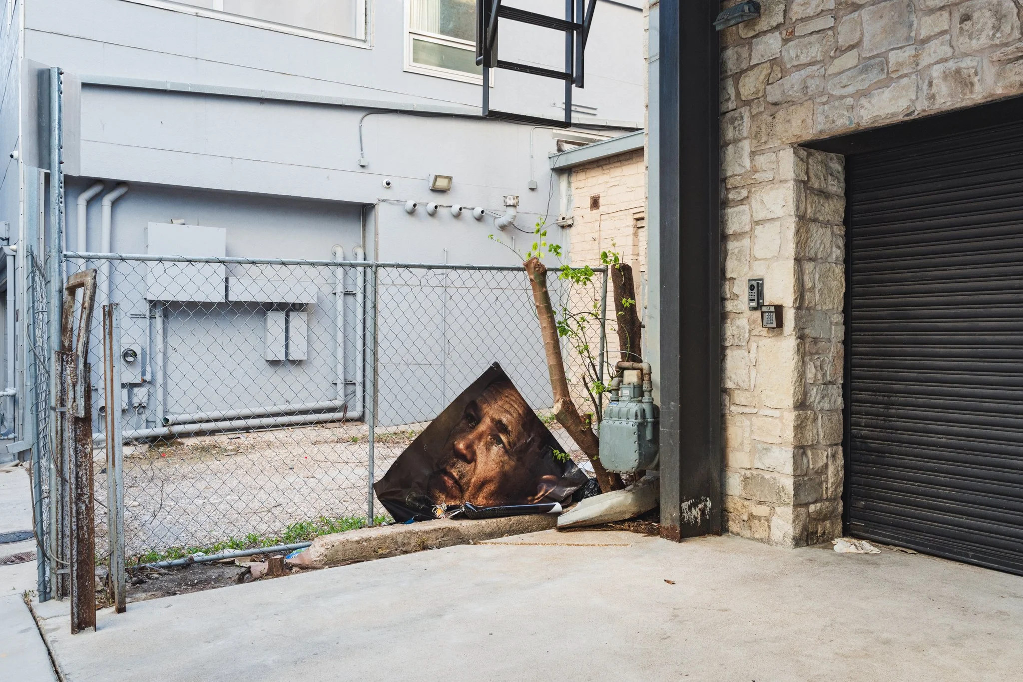 An alleyway with a chain-link fence, discarded poster of a man's face, a small tree, and an electrical box, adjacent to a building with stone and metal siding.