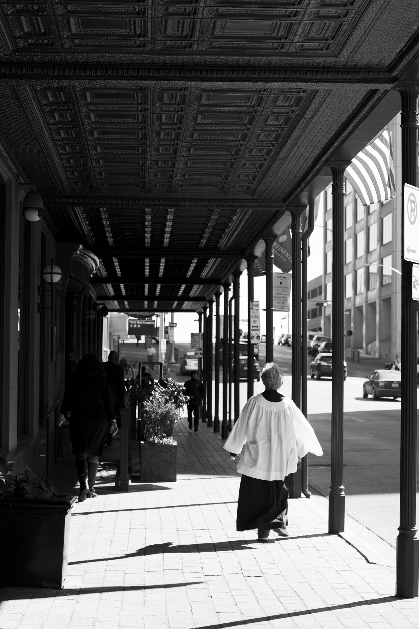 A black-and-white photo of a person wearing a white religious robe walking on a city sidewalk underneath a decorative overhead canopy, with other pedestrians and parked cars in the background.