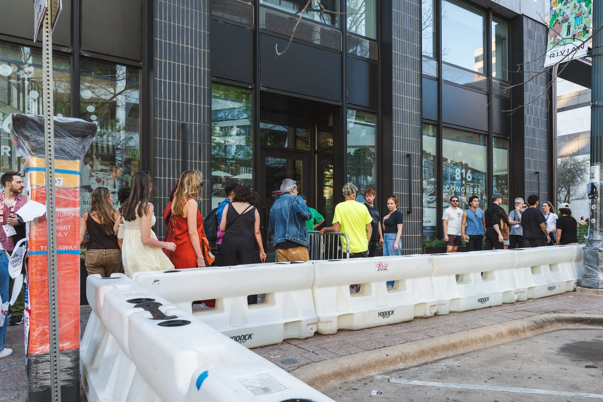 People gathered outside a modern black building with glass windows, some talking, in an urban setting with a barricade in the foreground.