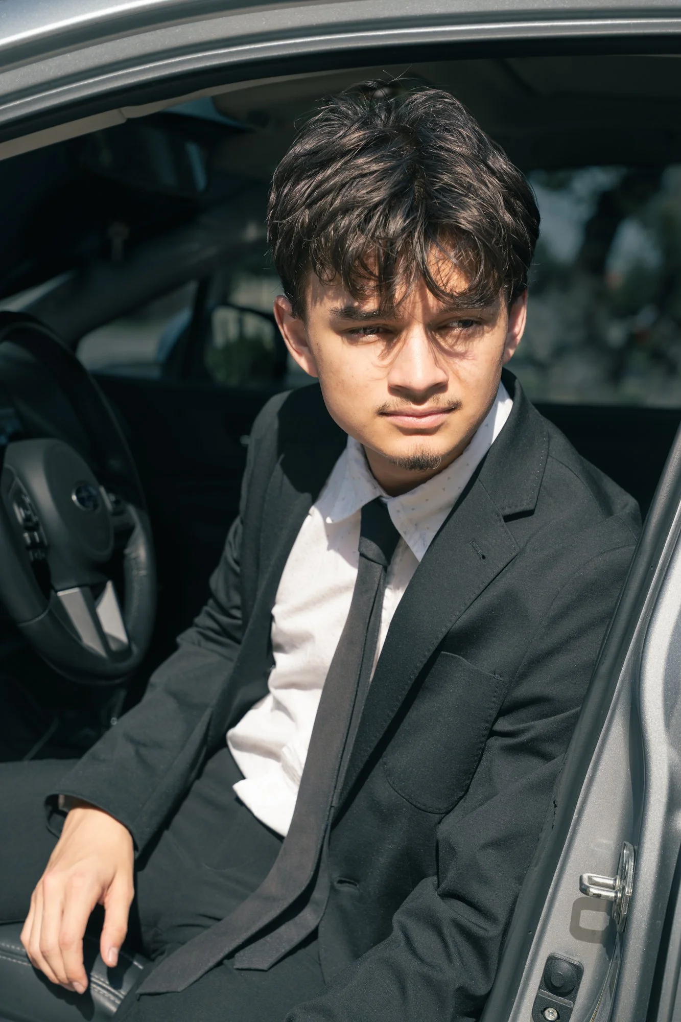 Young man in a black suit and tie sitting in the driver's seat of a car, looking out through the open car door with a serious expression.