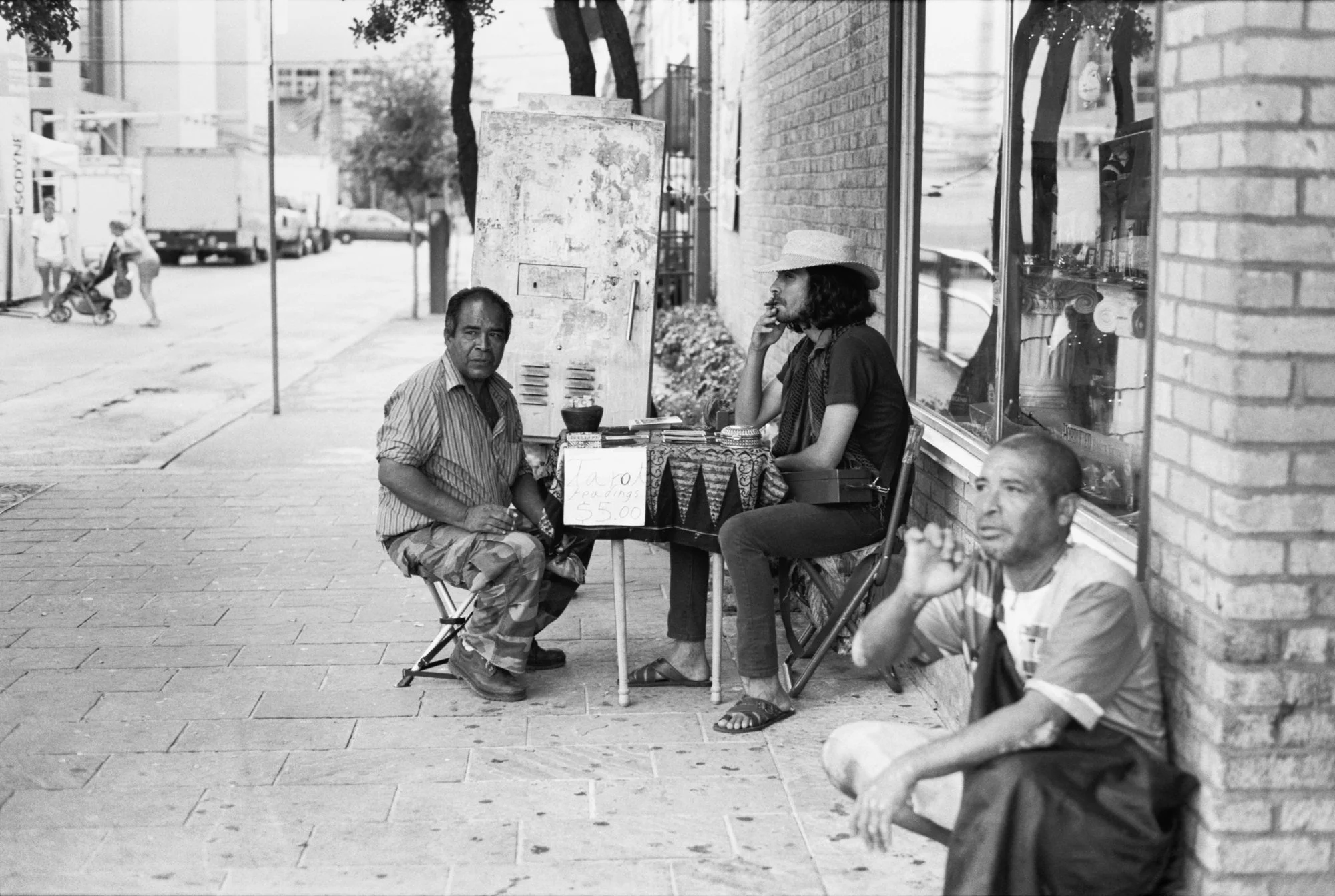 Three men sitting and standing on a sidewalk outside a store, with one man holding a cigarette. One man is seated on a small stool, another is sitting on the ground, and a third is seated on a chair. A table with a sign offering readings for $5 is in