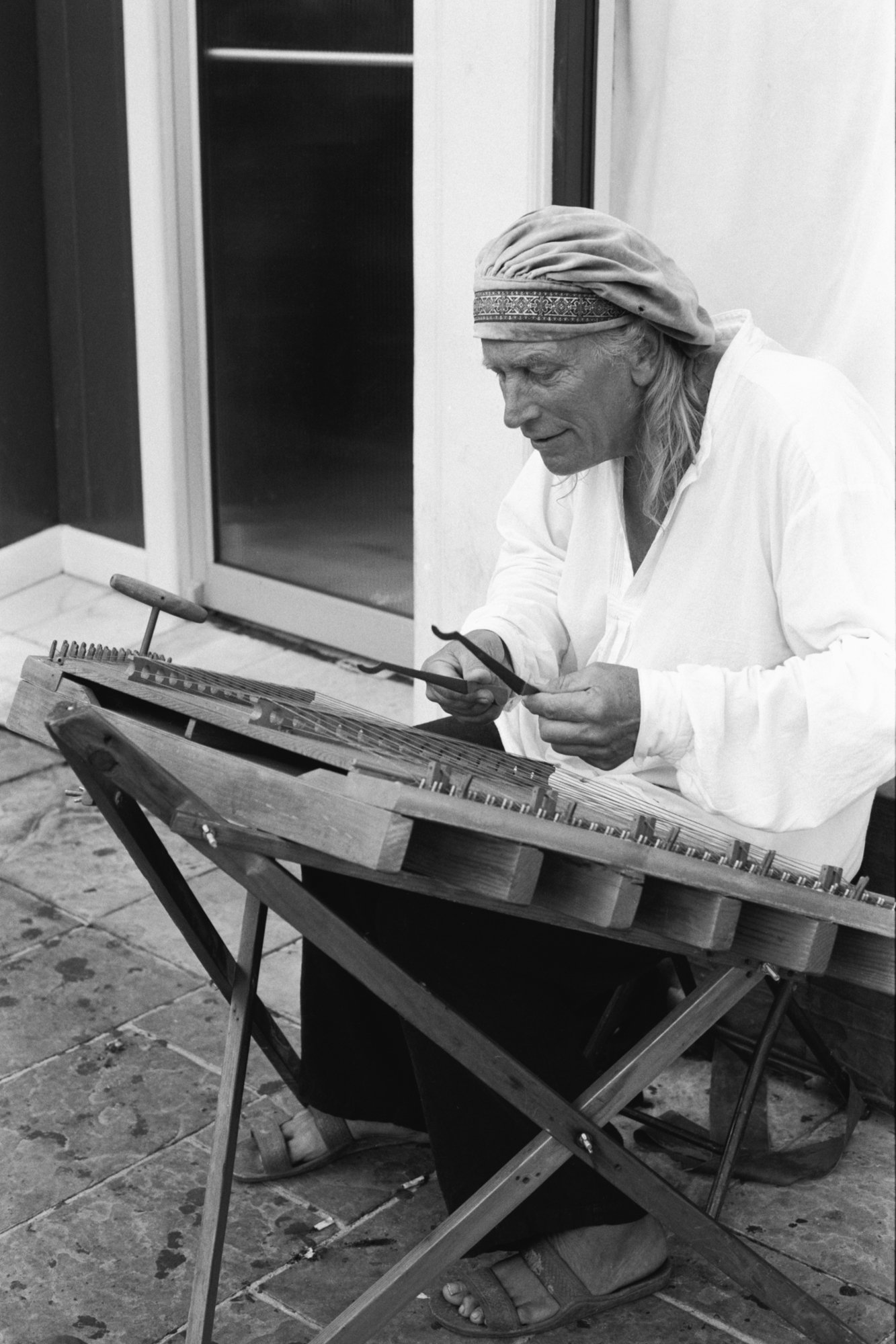 A man with long hair wearing a headband and a white shirt, sitting outdoors and playing a hammered dulcimer.