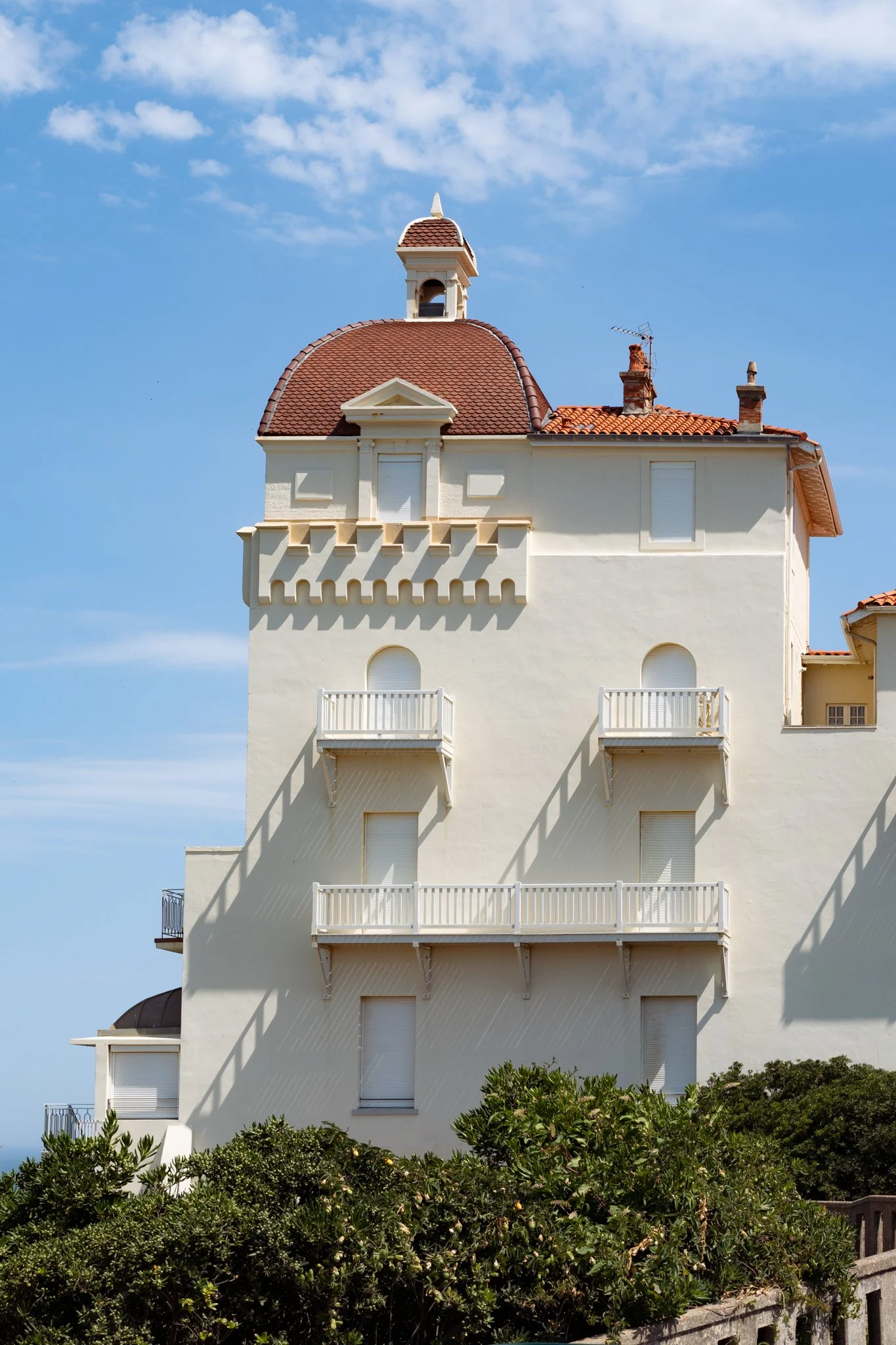 A multi-story white building with small balconies, arched windows, and a red-tiled roof. There are bushes at the bottom and a blue sky with clouds above.