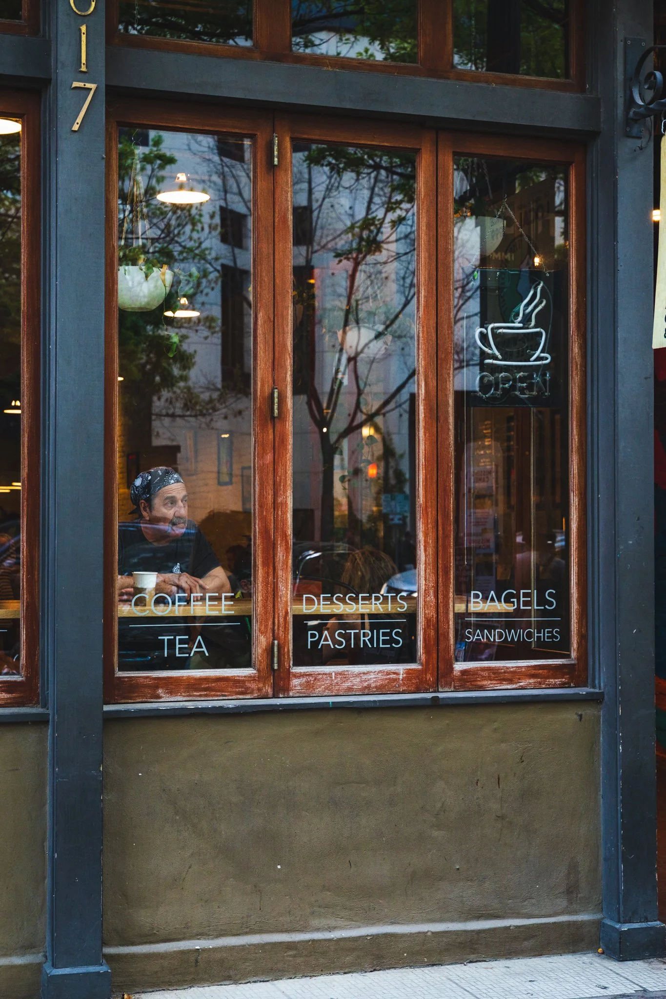 The window of a cafe with a neon sign saying 'Open'. Inside, a person with a bandana is sitting at a table with a cup. The window has words like 'Coffee', 'Tea', 'Desserts', 'Pastries', 'Bagels', and 'Sandwiches' written on the glass.