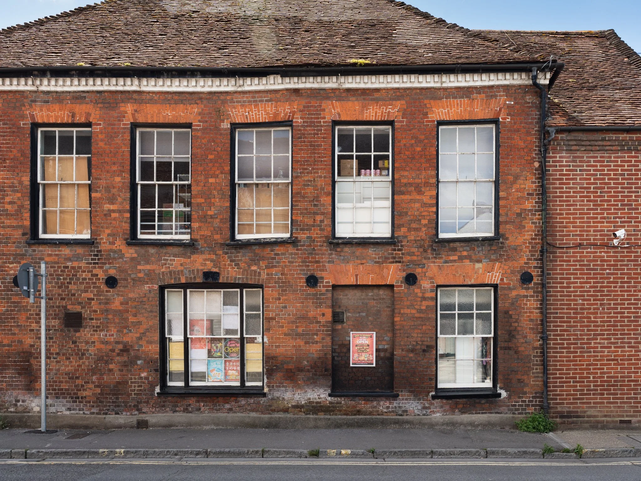 A brick building with four large windows and a storefront entrance, featuring signs and posters in the windows, a streetlamp, and a sidewalk in front.