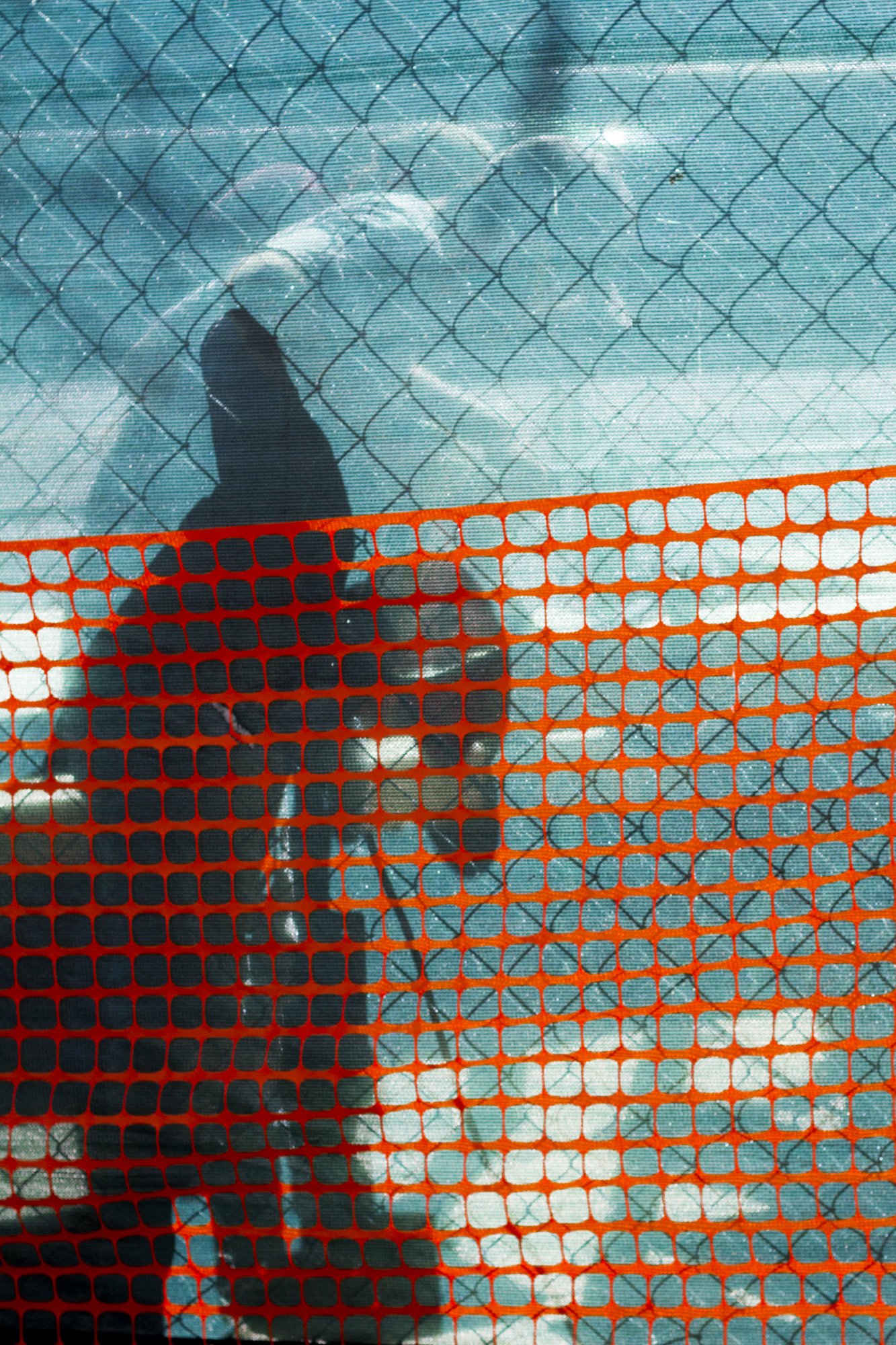 A person wearing black shoes and black clothing walking on a snowy or icy surface, viewed through a chain-link fence with orange plastic snow fence in the foreground.
